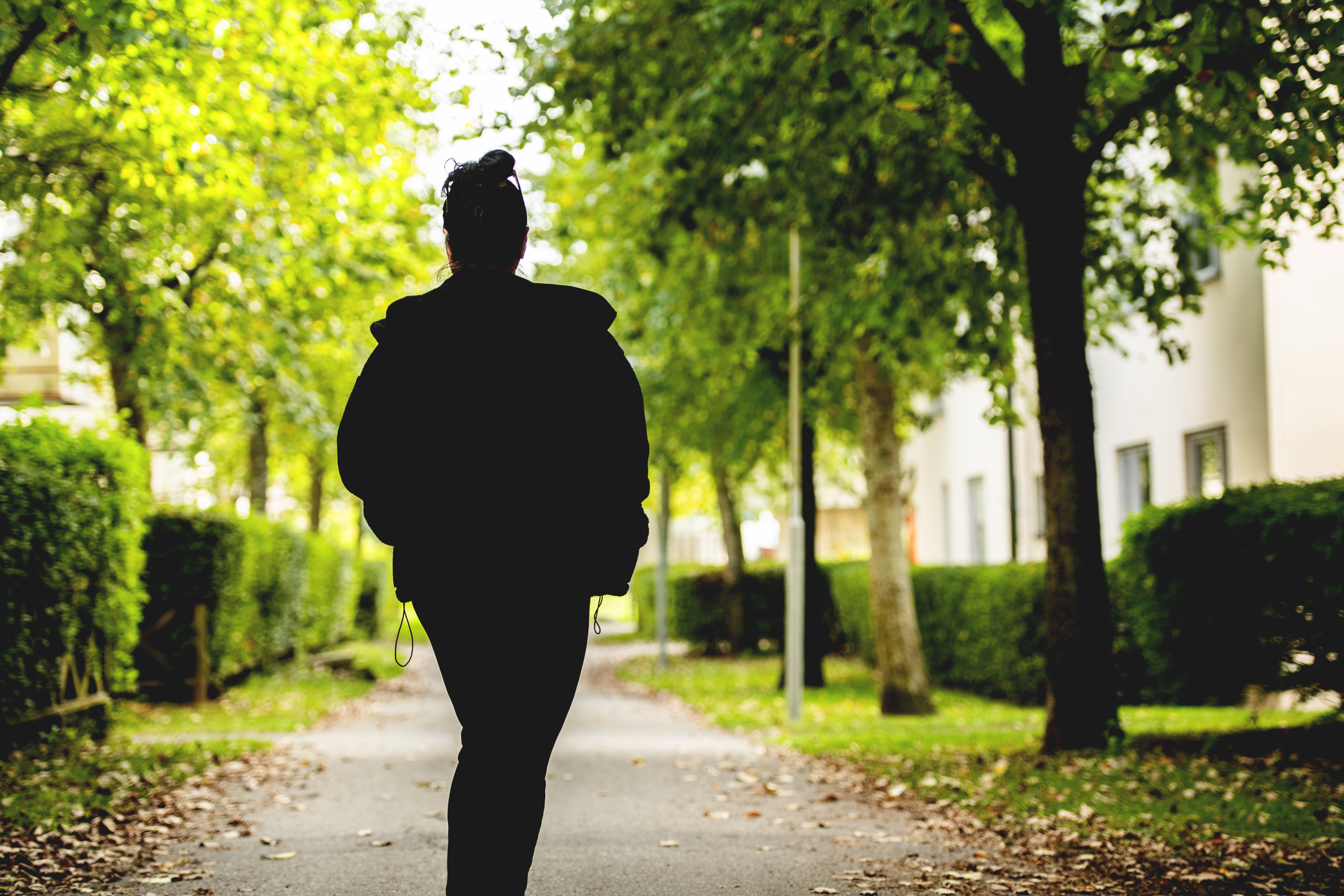 Leyla i mörka kläder går ensam på en väg med träd i en park eller ett bostadsområde, omgiven av växter och löv.
