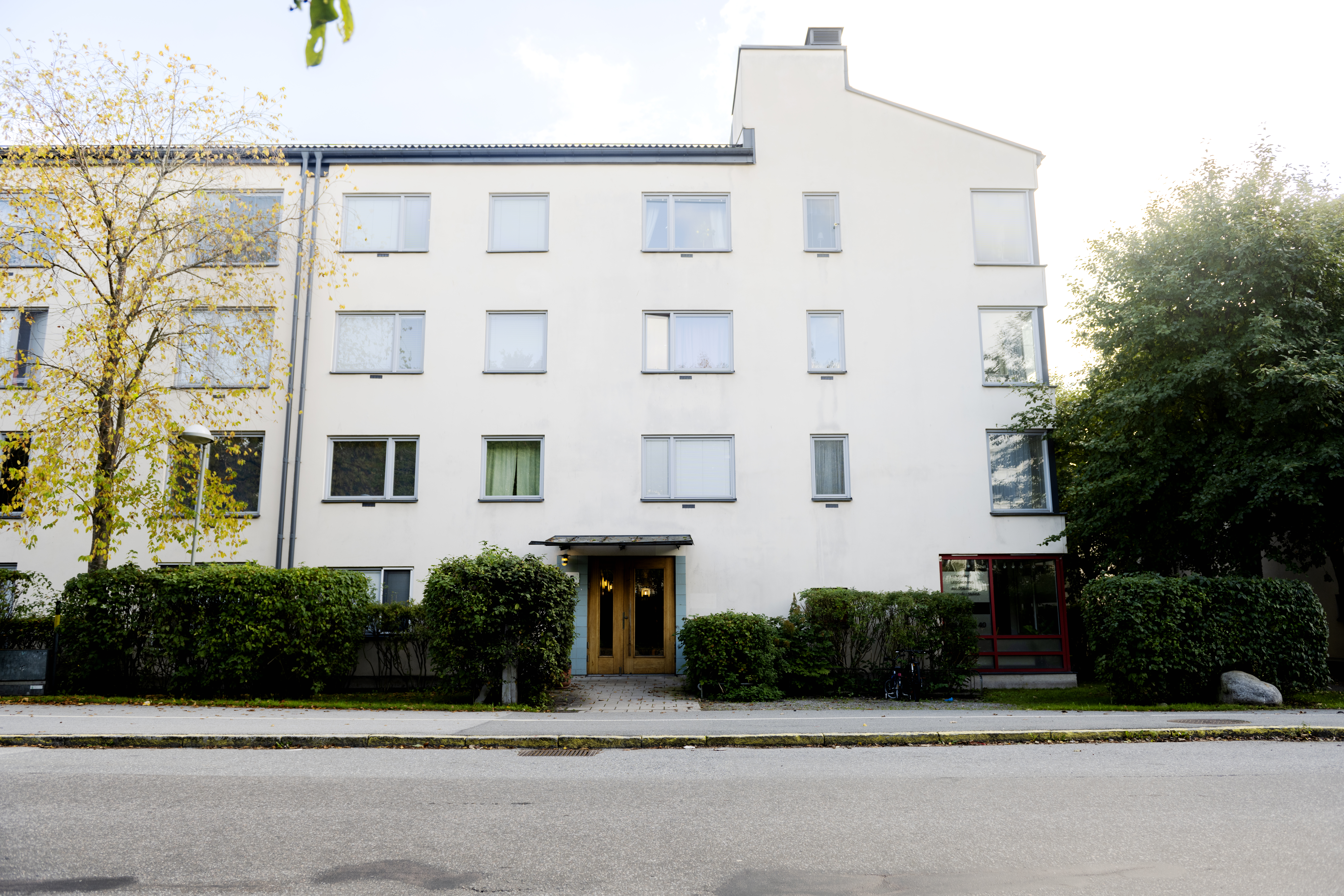 Ett fyravånings vitt lägenhetshus med flera fönster, en centralt placerad ingång och buskar framför, sett från gatan.