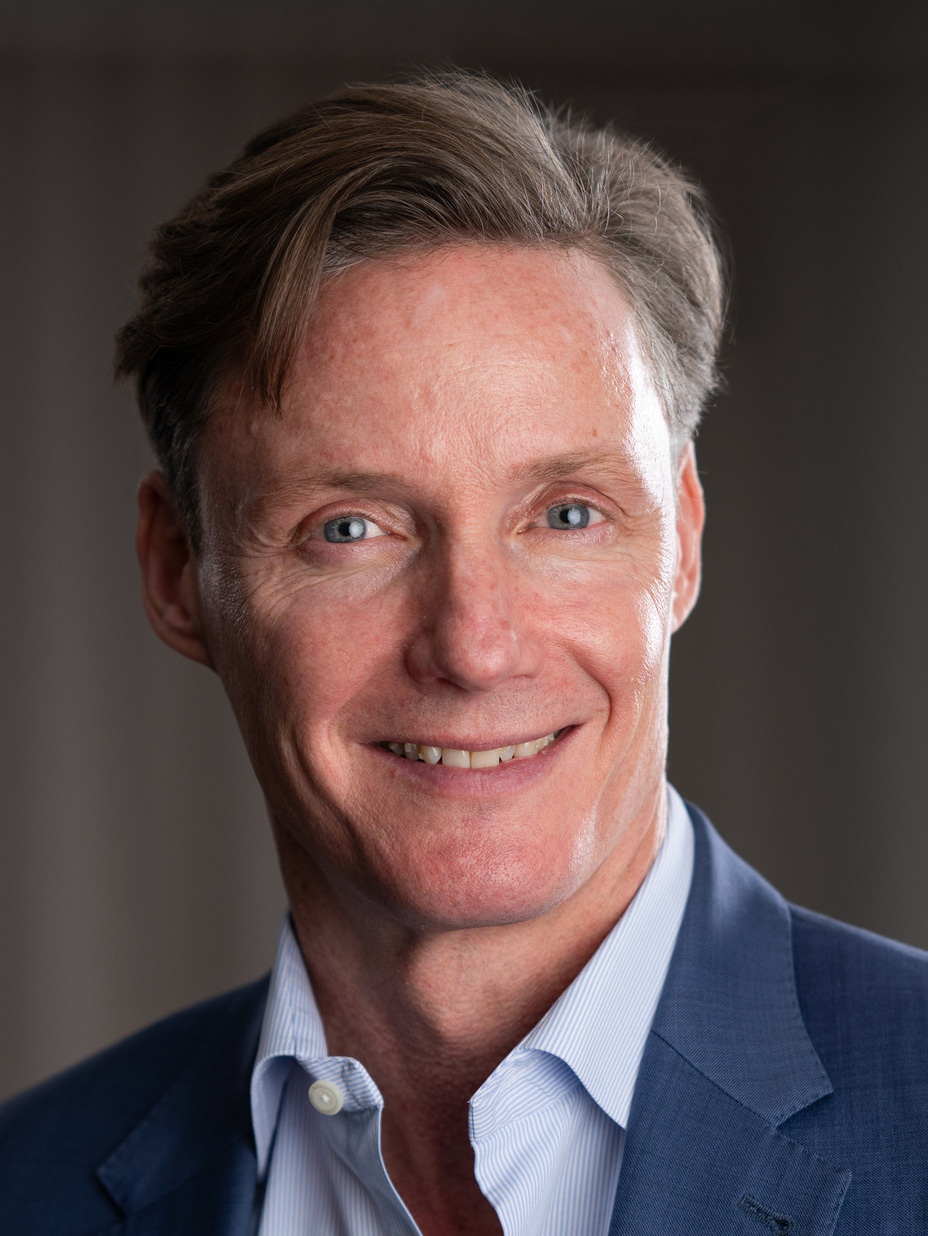 En medelålders man med ljus hud och kort brunt hår, iklädd blå kavaj och ljusblå skjorta, ler mot kameran mot neutral bakgrund.