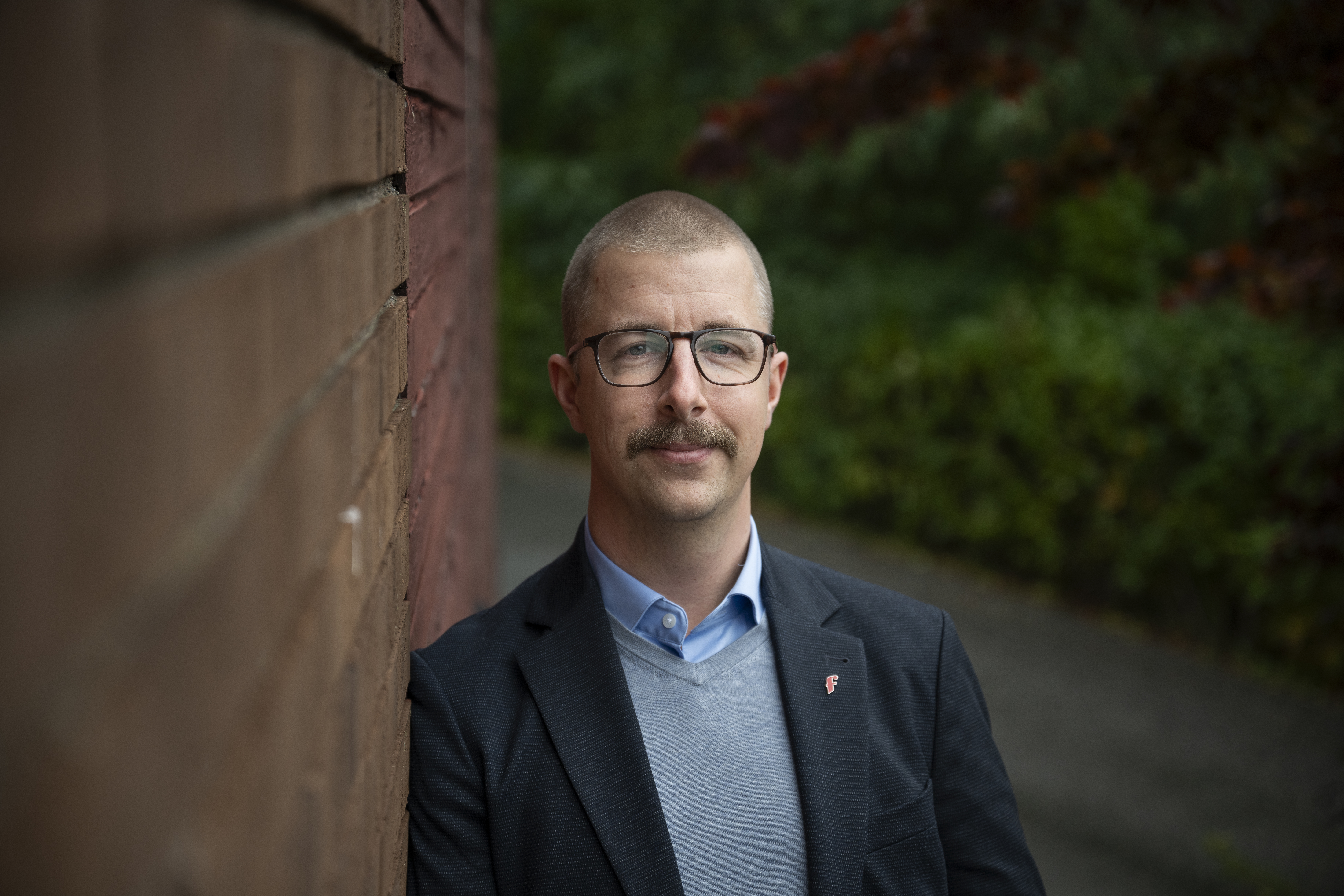 Bild på man i glasögon och kavaj vid en tegelvägg.