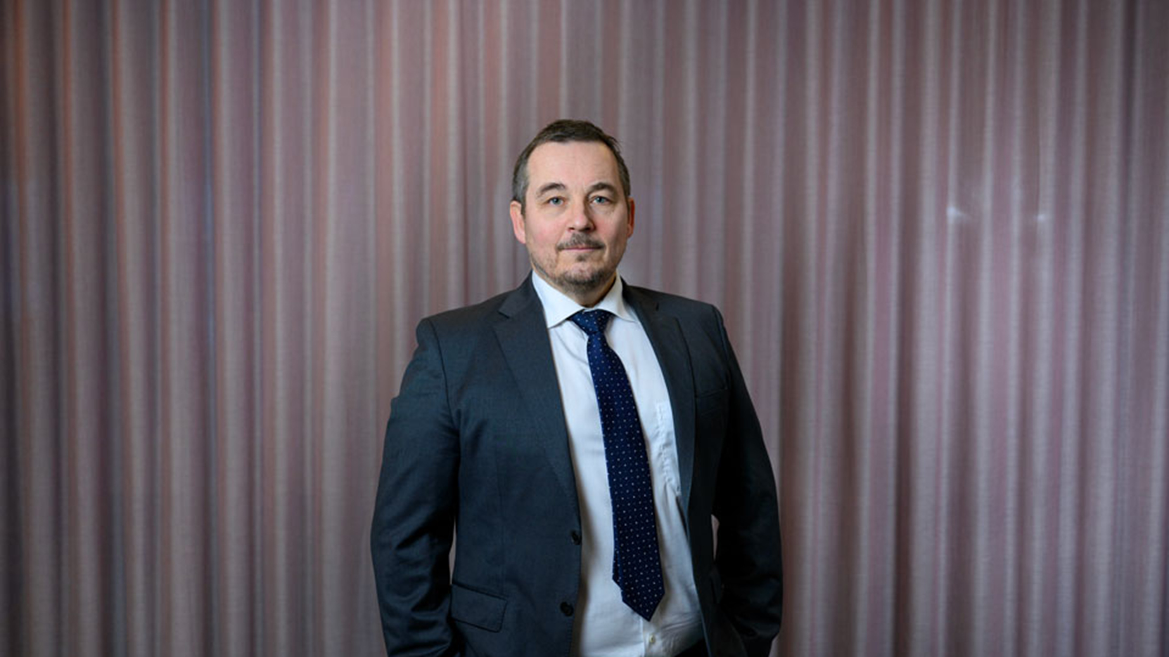 En man i kostym och randig slips står framför ett rosa draperi och tittar in i kameran med neutral min.