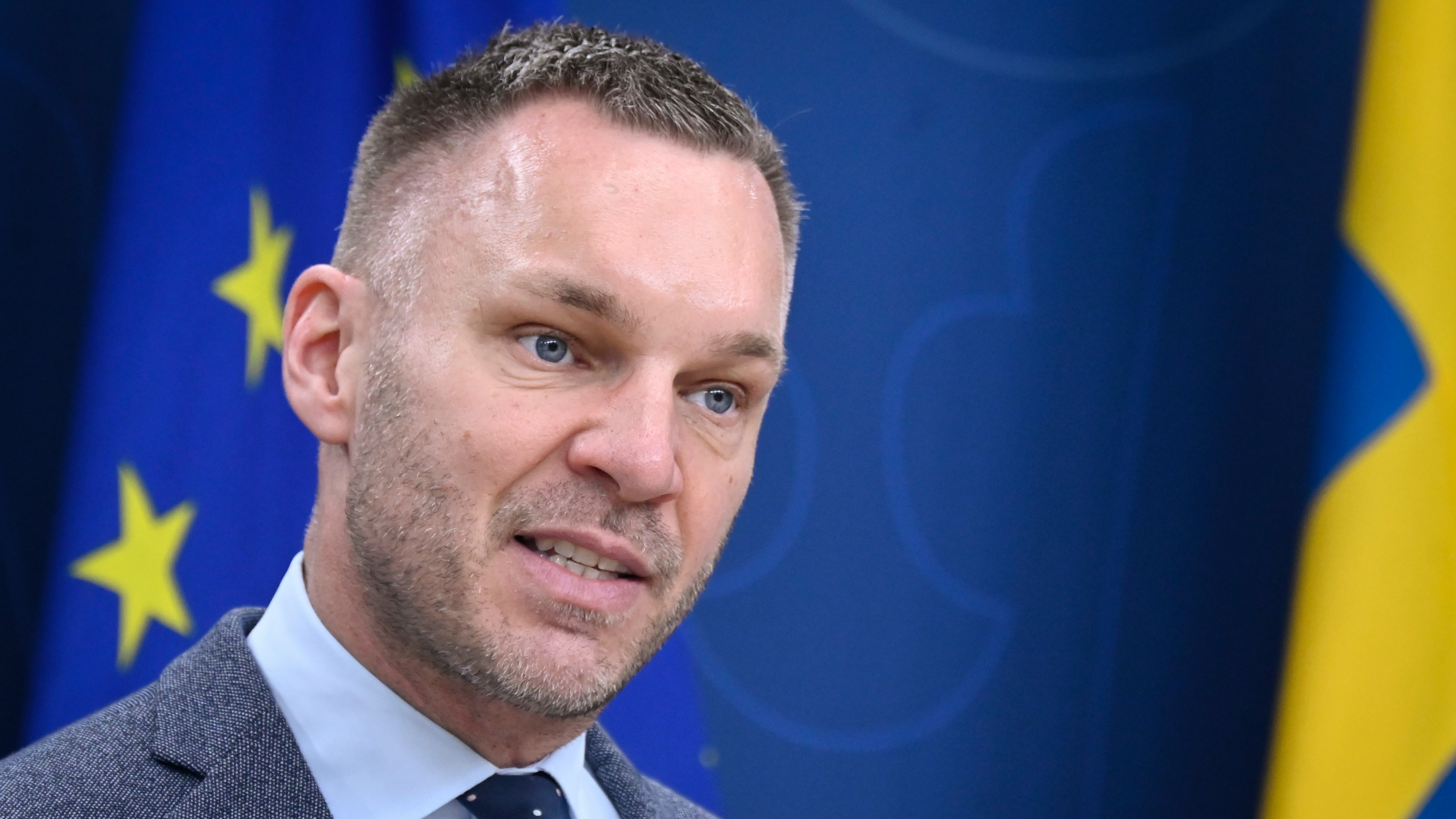 Erik Slottner i kostym talar framför blå flaggor med gula stjärnor och en gul form, vilket antyder ett europeiskt sammanhang.