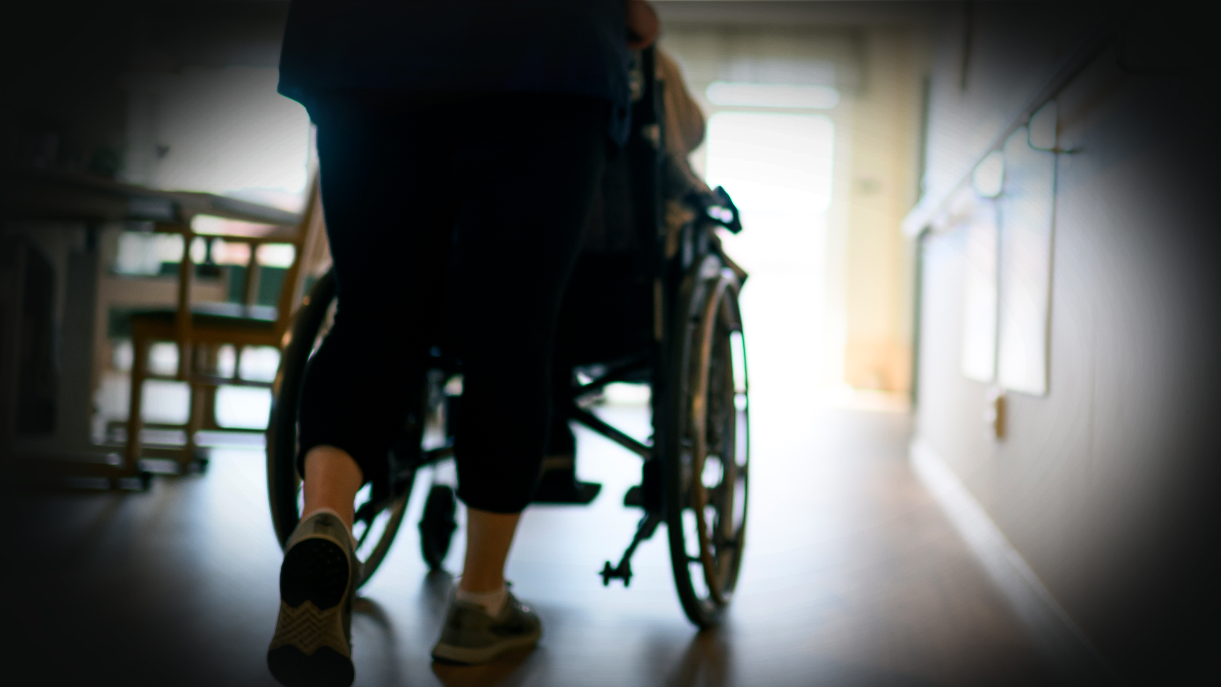 En person skjuter någon i rullstol genom en korridor, med ljus från ett fönster i bakgrunden.