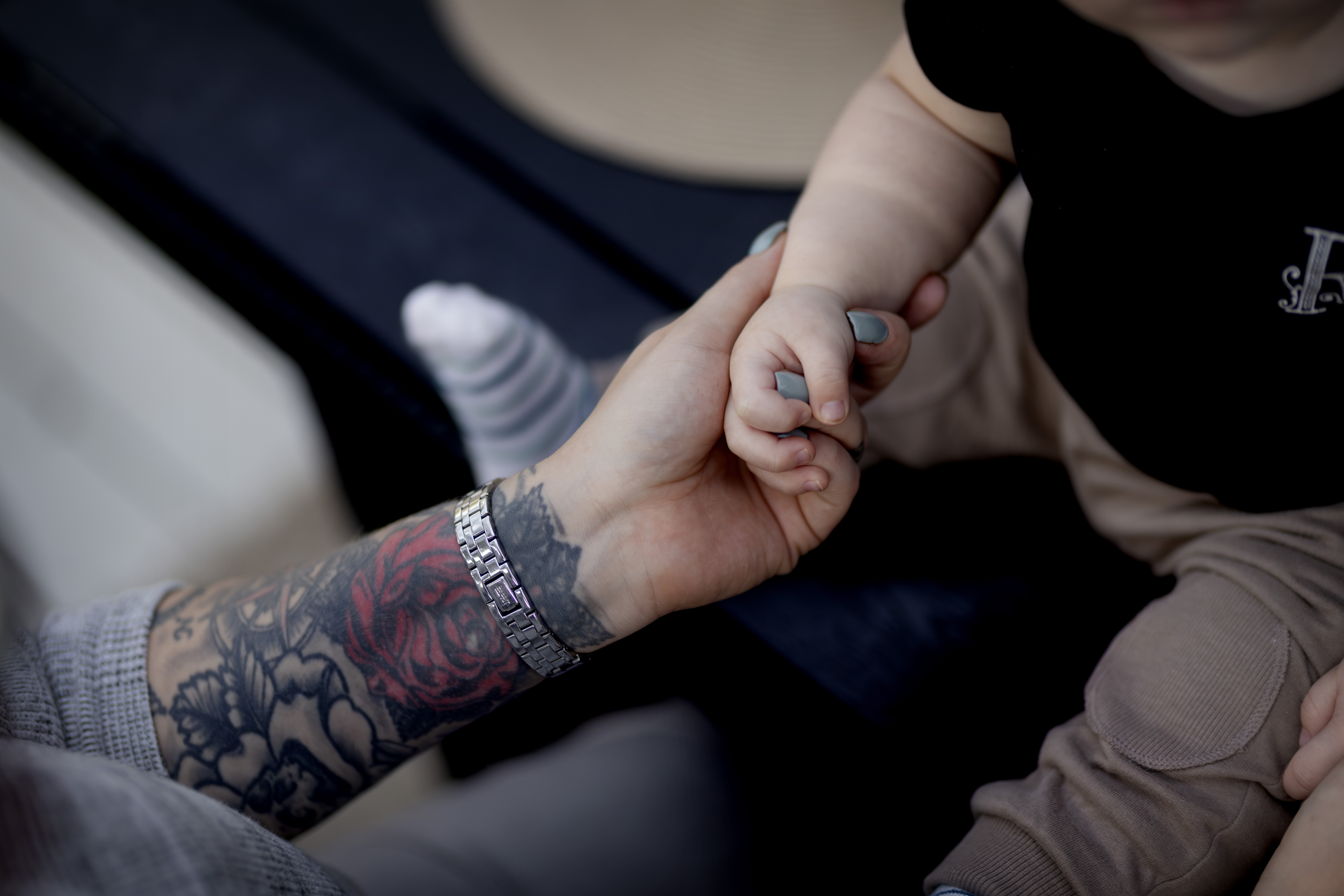 En vuxen med tatuerad arm och armbandsur håller en bebis i handen. Bebisen sitter och har ljusa byxor samt svart tröja.