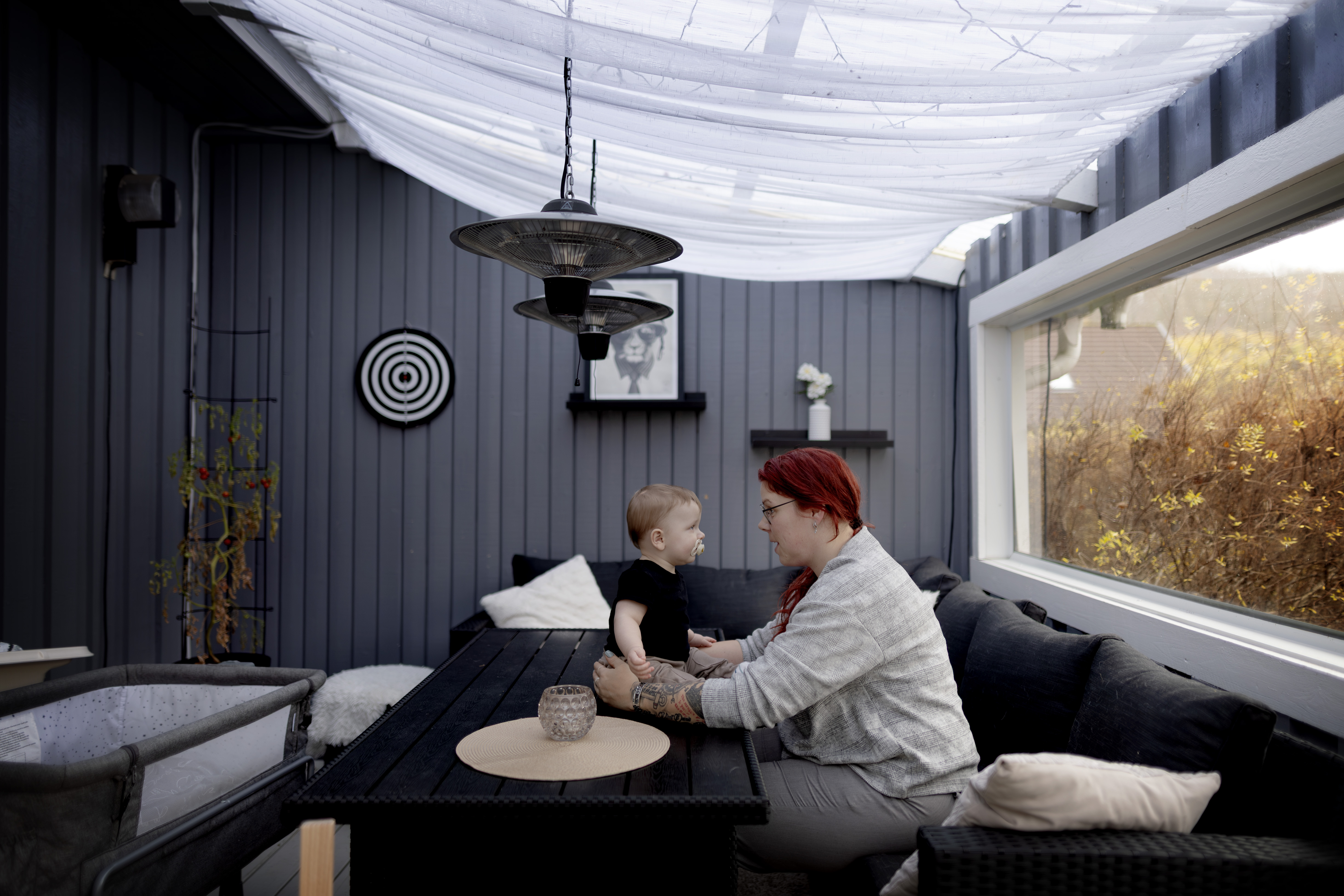 En vuxen med rött hår sitter vid ett svart bord mittemot ett litet barn på en täckt uteplats.