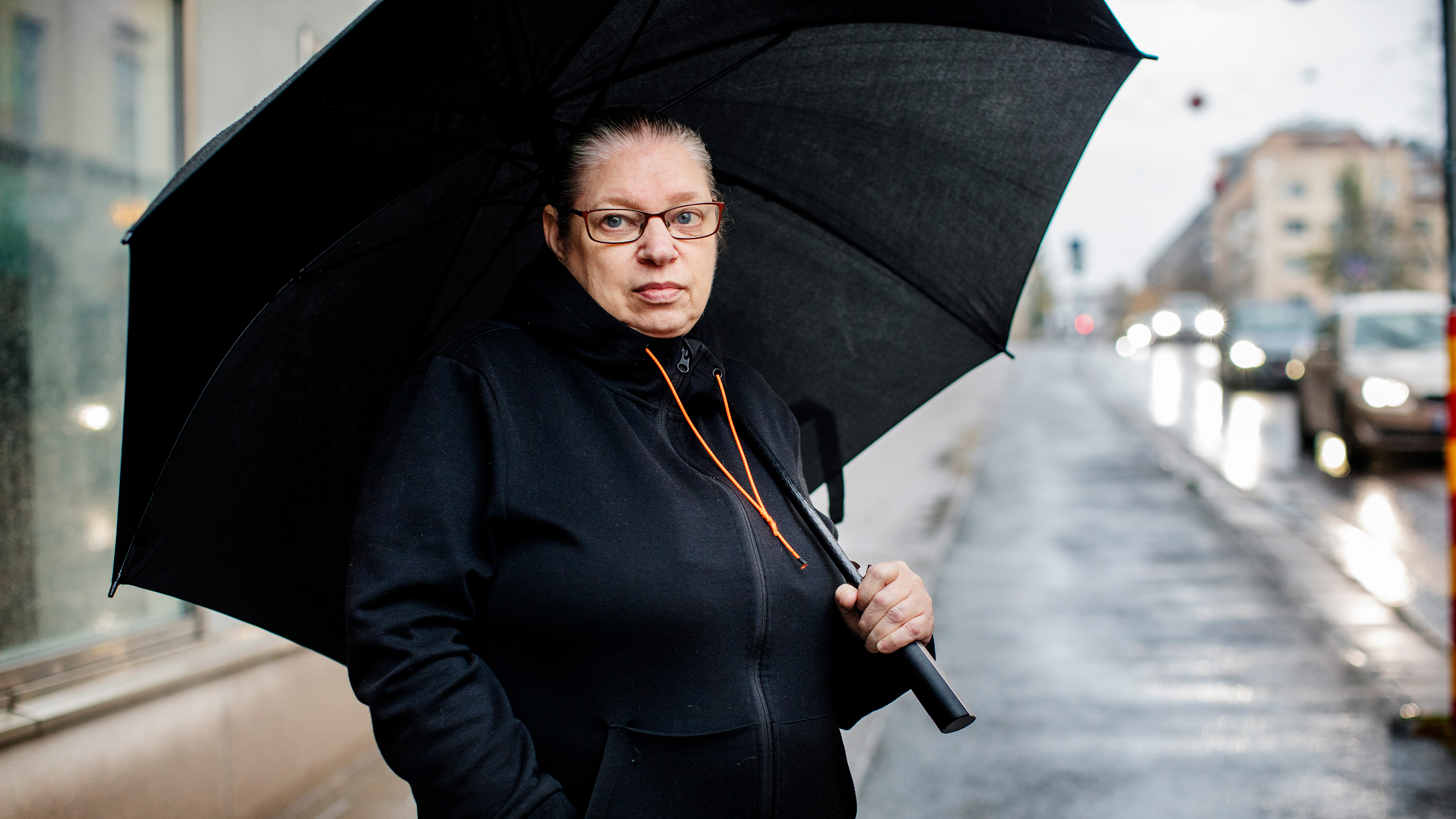 Marita Rollin i glasögon och svart jacka står på en trottoar i en stad och håller ett svart paraply under regn.