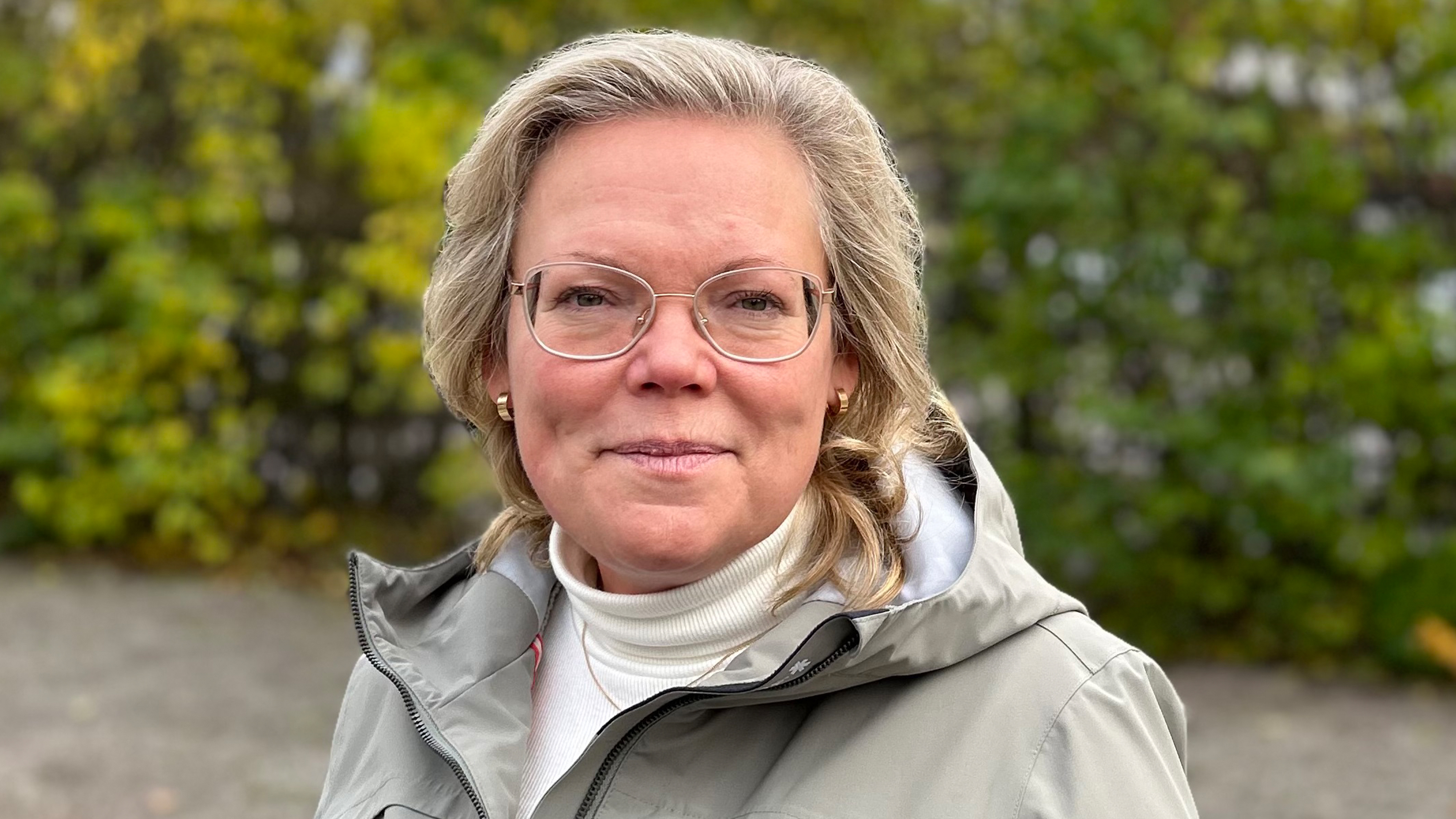 En kvinna med blont hår och glasögon har en grå jacka och vit polotröja och står utomhus framför grönska.