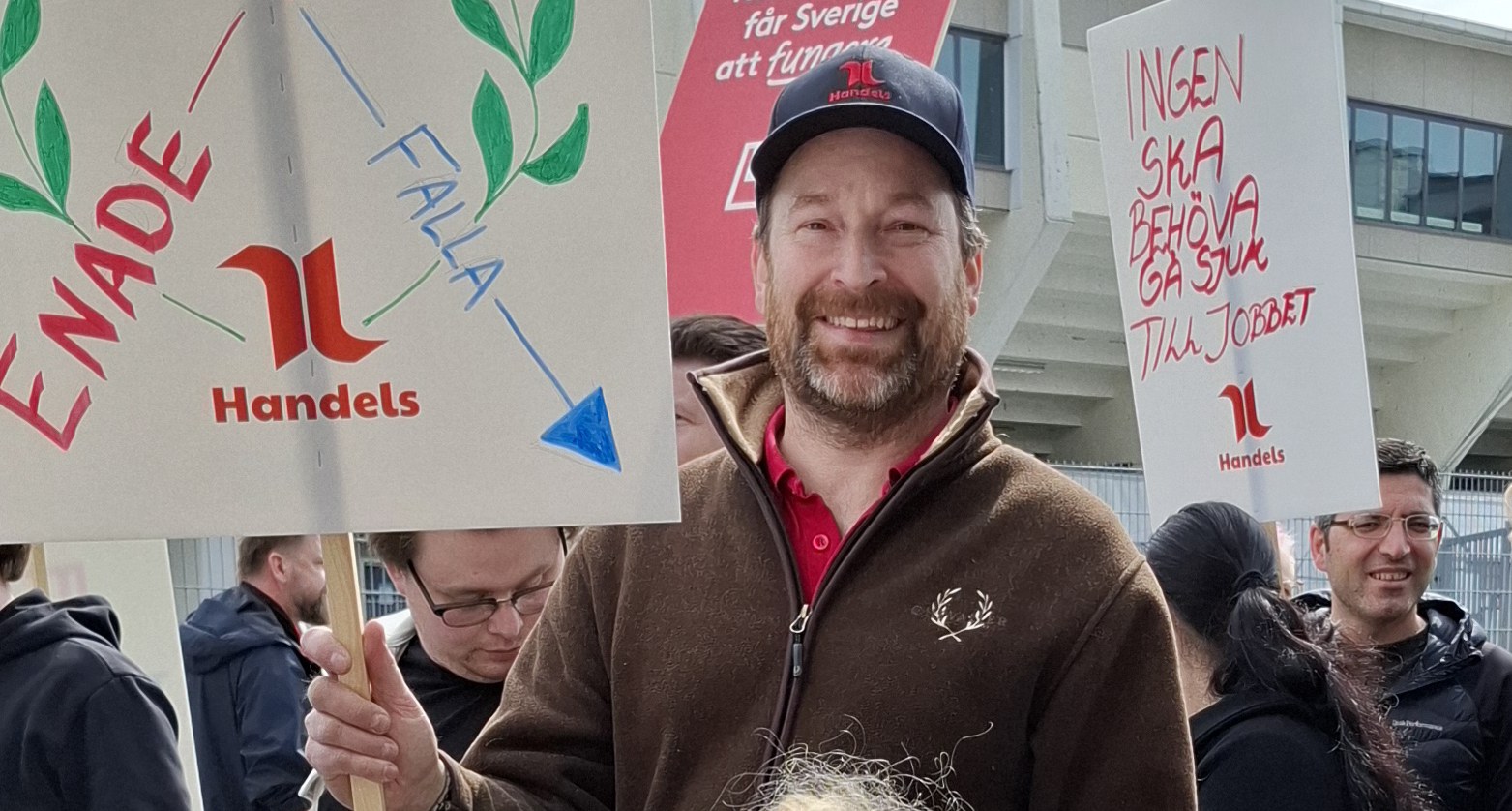 En man i röd tröja och keps håller ett Handels-plakat; andra personer med skyltar syns i bakgrunden.