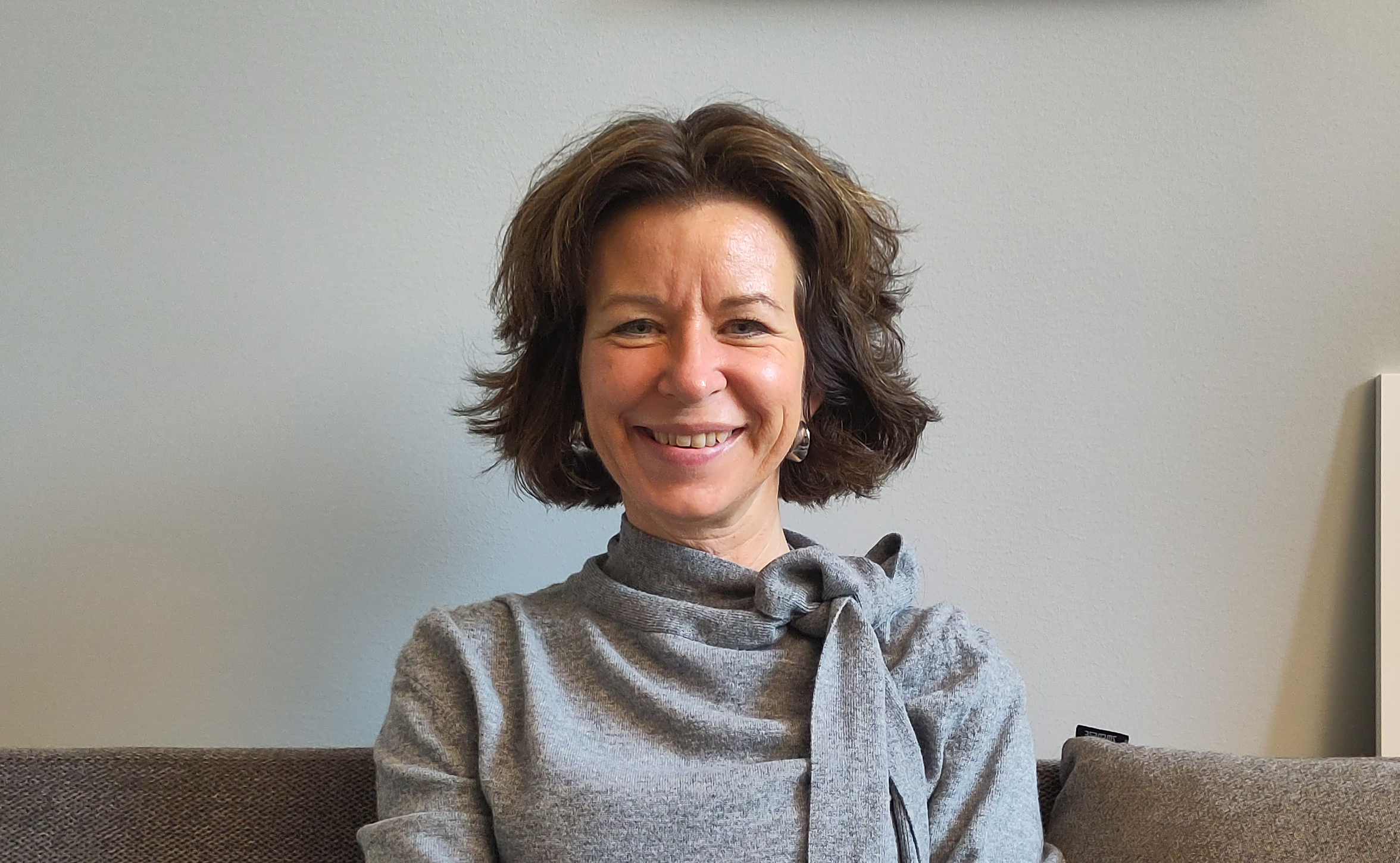 En kvinna med kort brunt hår har en grå topp med rosett, sitter i en soffa och ler mot kameran framför en ljus vägg.