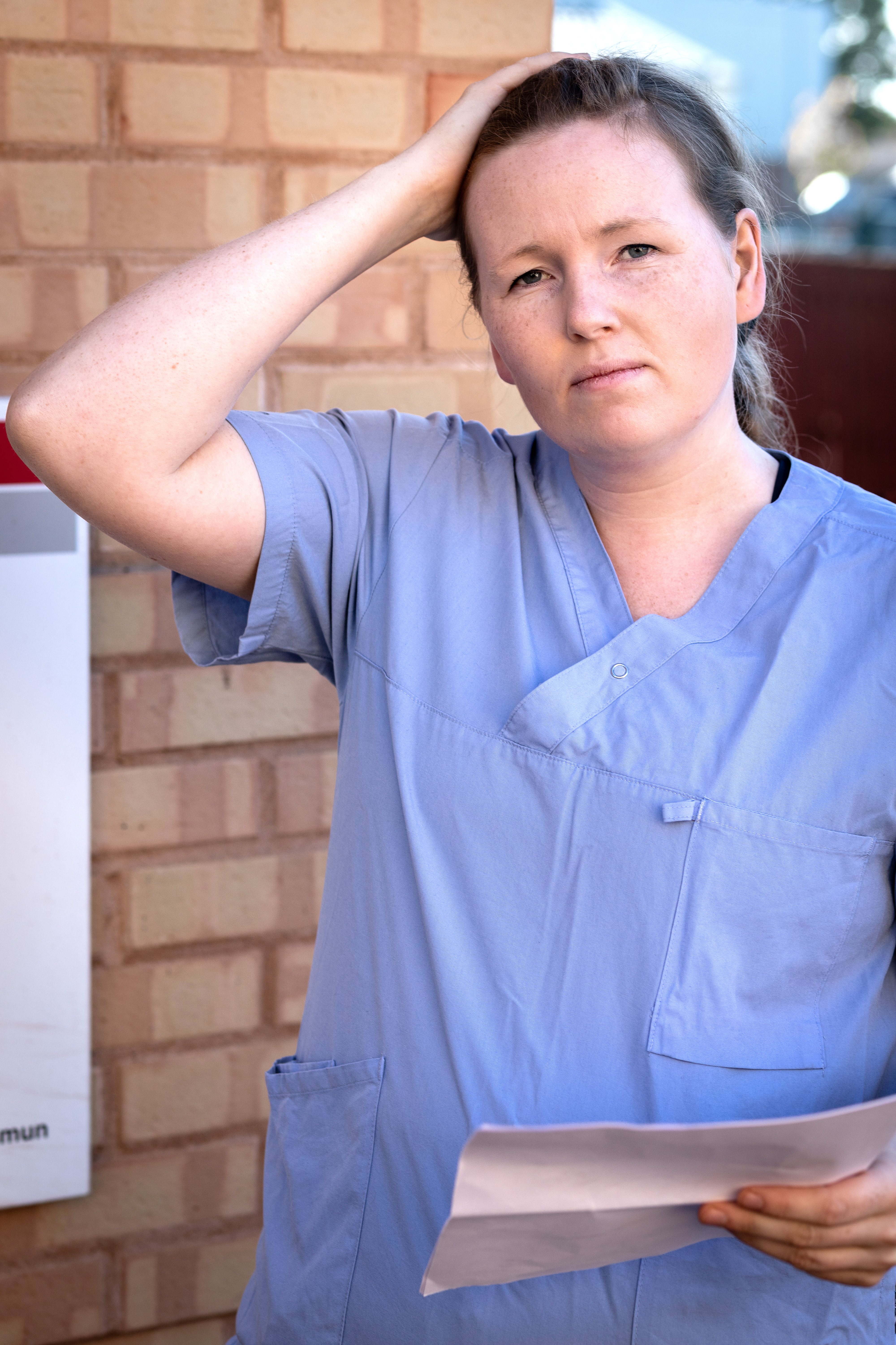 En person i medicinska kläder står utomhus, håller ett papper och rör vid sitt huvud.