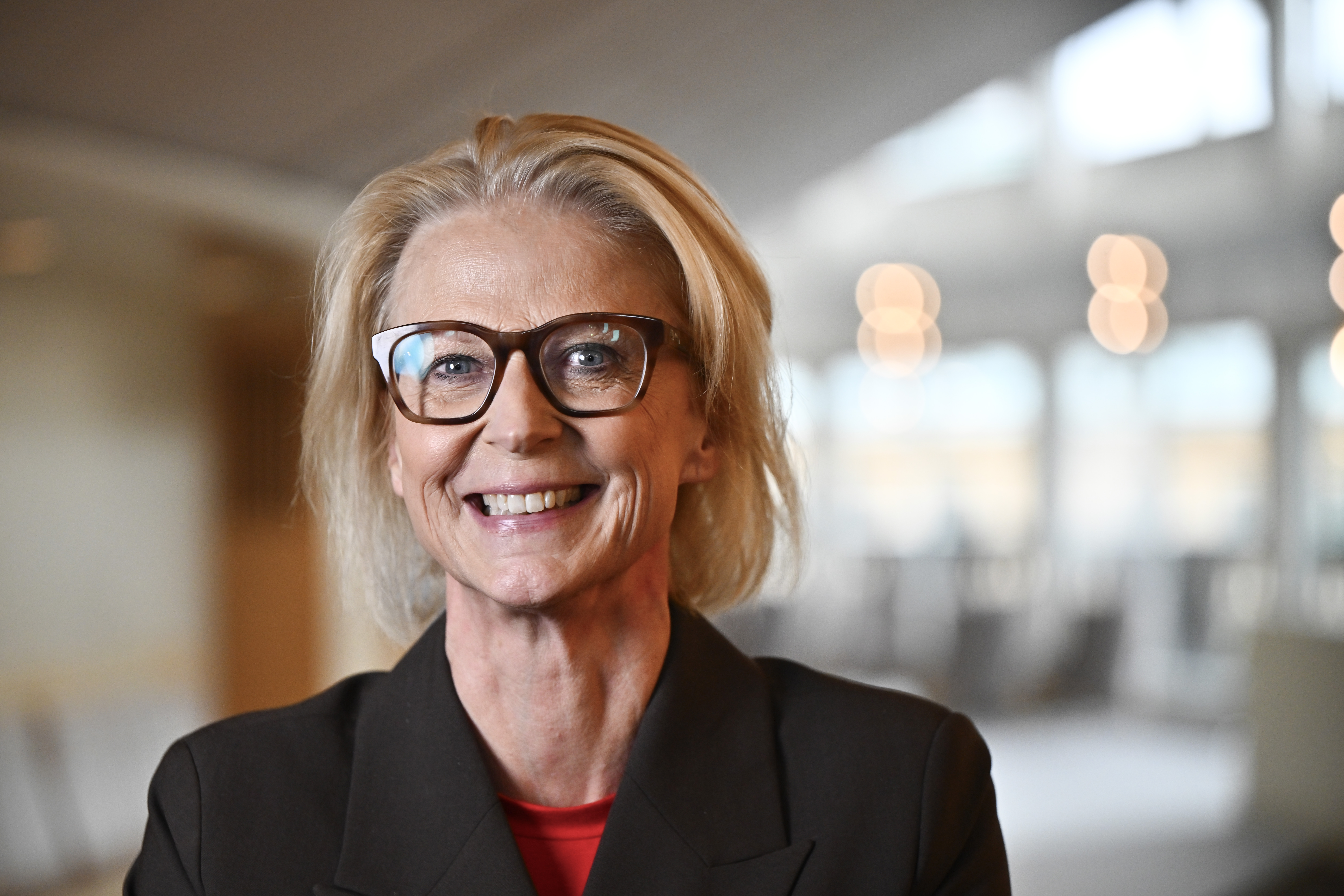Elisabeth Svantesson blont hår och glasögon har på sig en mörk kavaj och röd tröja och ler inomhus.