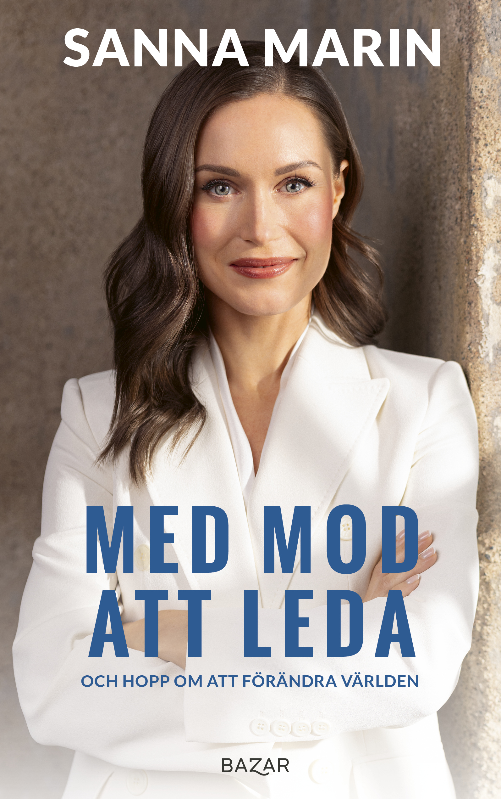 En kvinna med långt brunt hår i vit kostym står mot en vägg med armarna i kors, med texten "Med mod att leda" och "Sanna Marin".