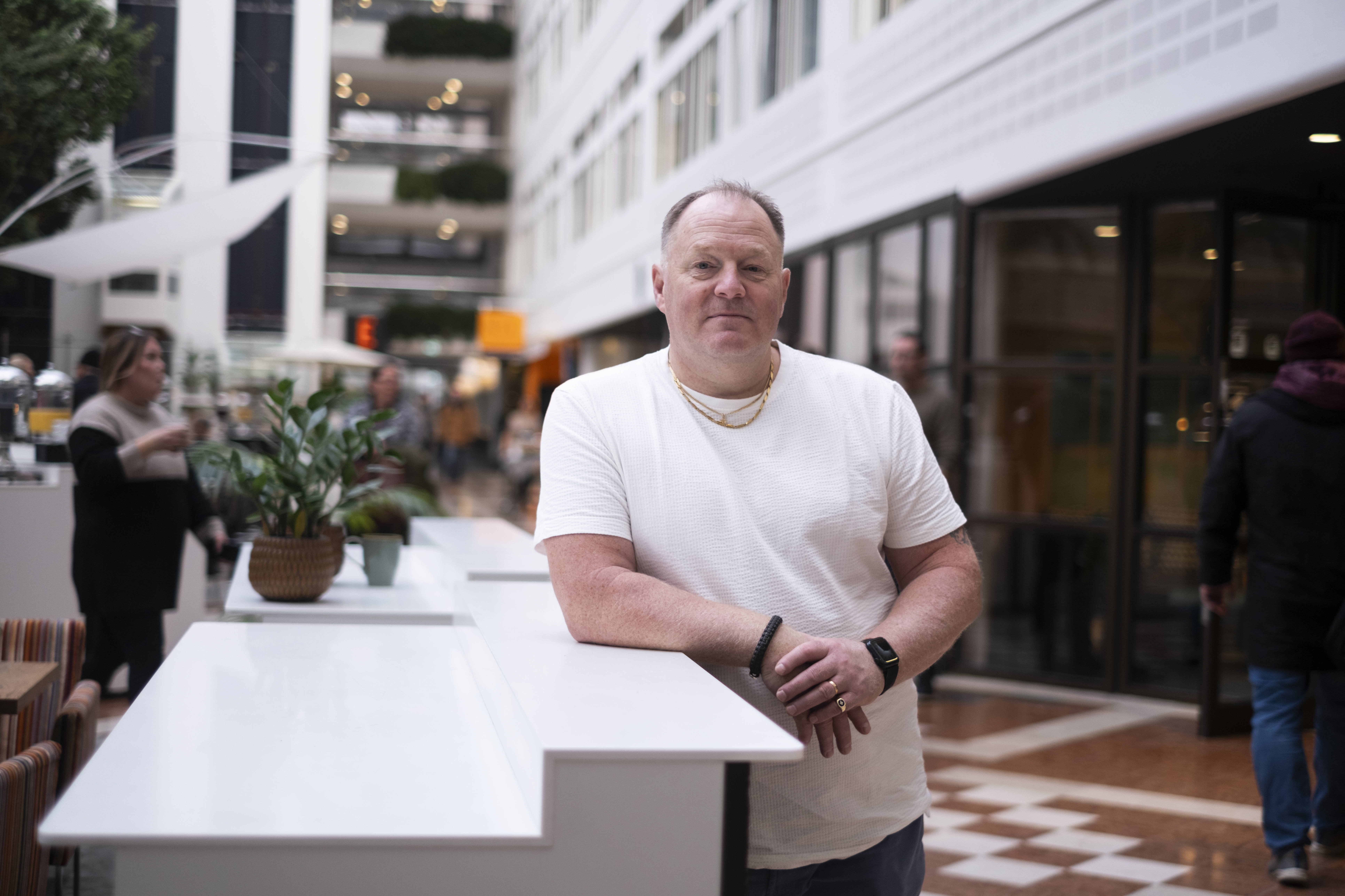 En man i vit t-shirt lutar sig mot en vit disk i en inomhuslokal med andra personer och växter i bakgrunden.