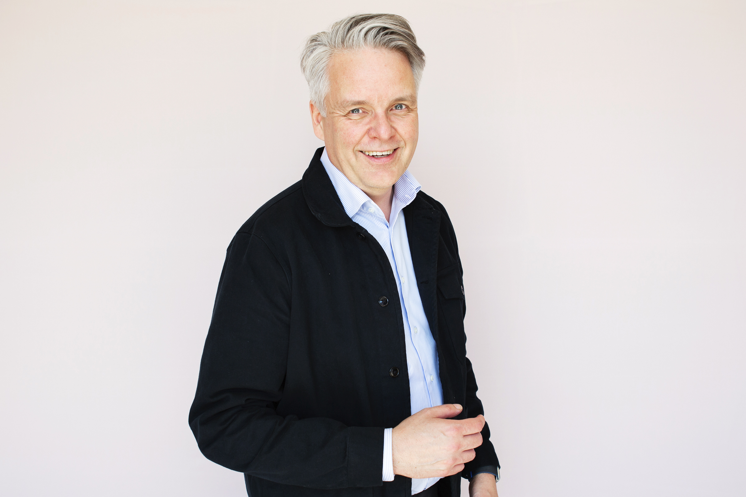 En man med grått hår har en svart jacka över en ljusblå skjorta och står mot en ljus bakgrund.