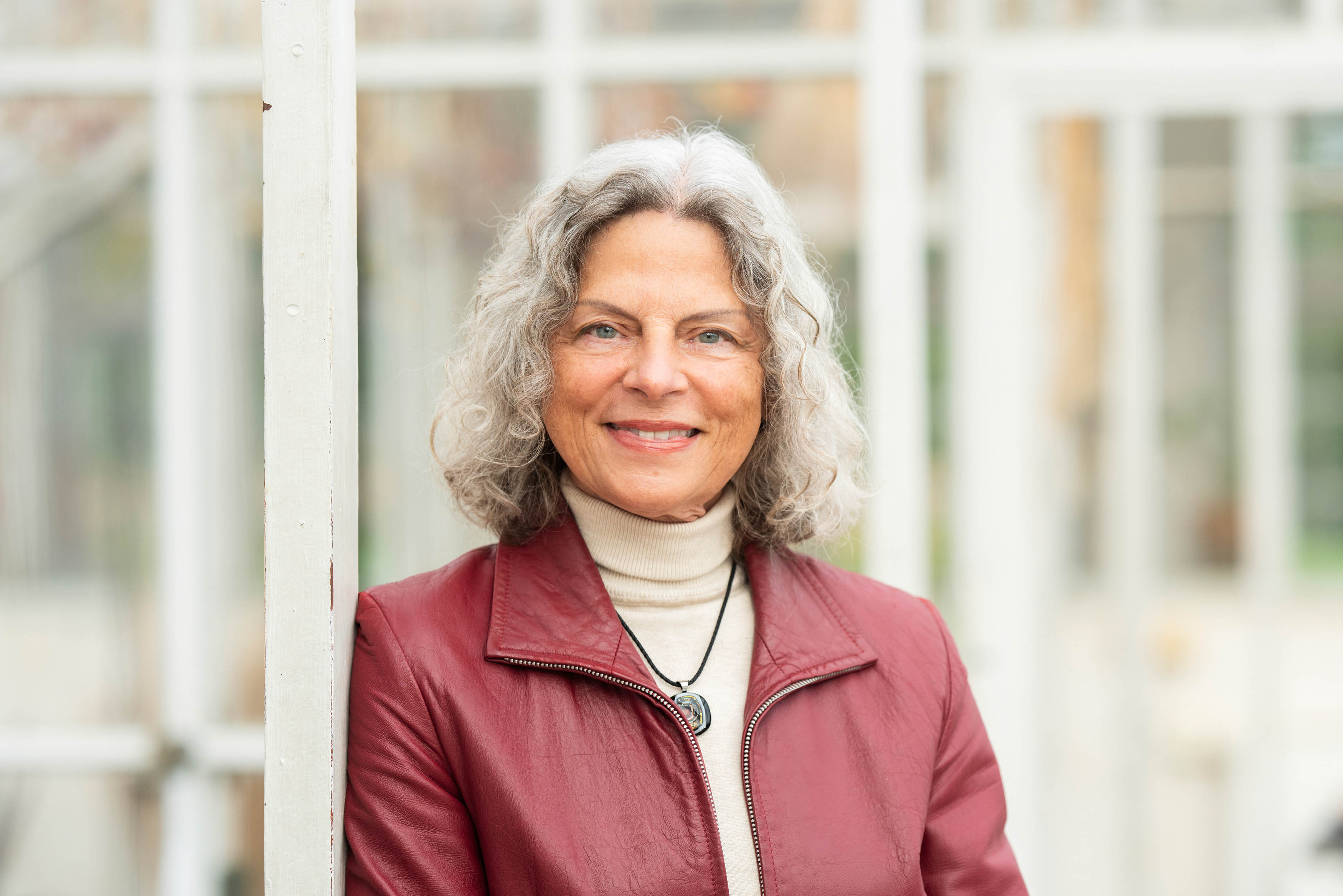 En kvinna med grått lockigt hår, iförd en mörkröd jacka och ljus polotröja, står inomhus och ler mot kameran.