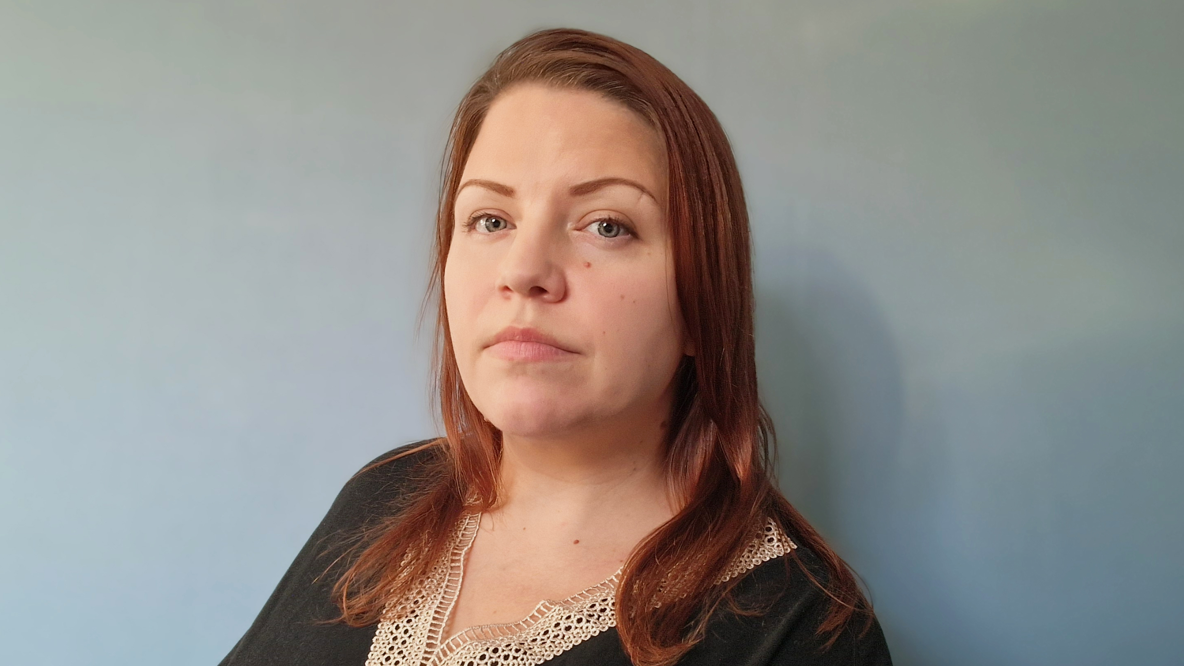 En kvinna med långt brunt hår i svart topp med beige spetskrage står framför en ljusblå bakgrund.