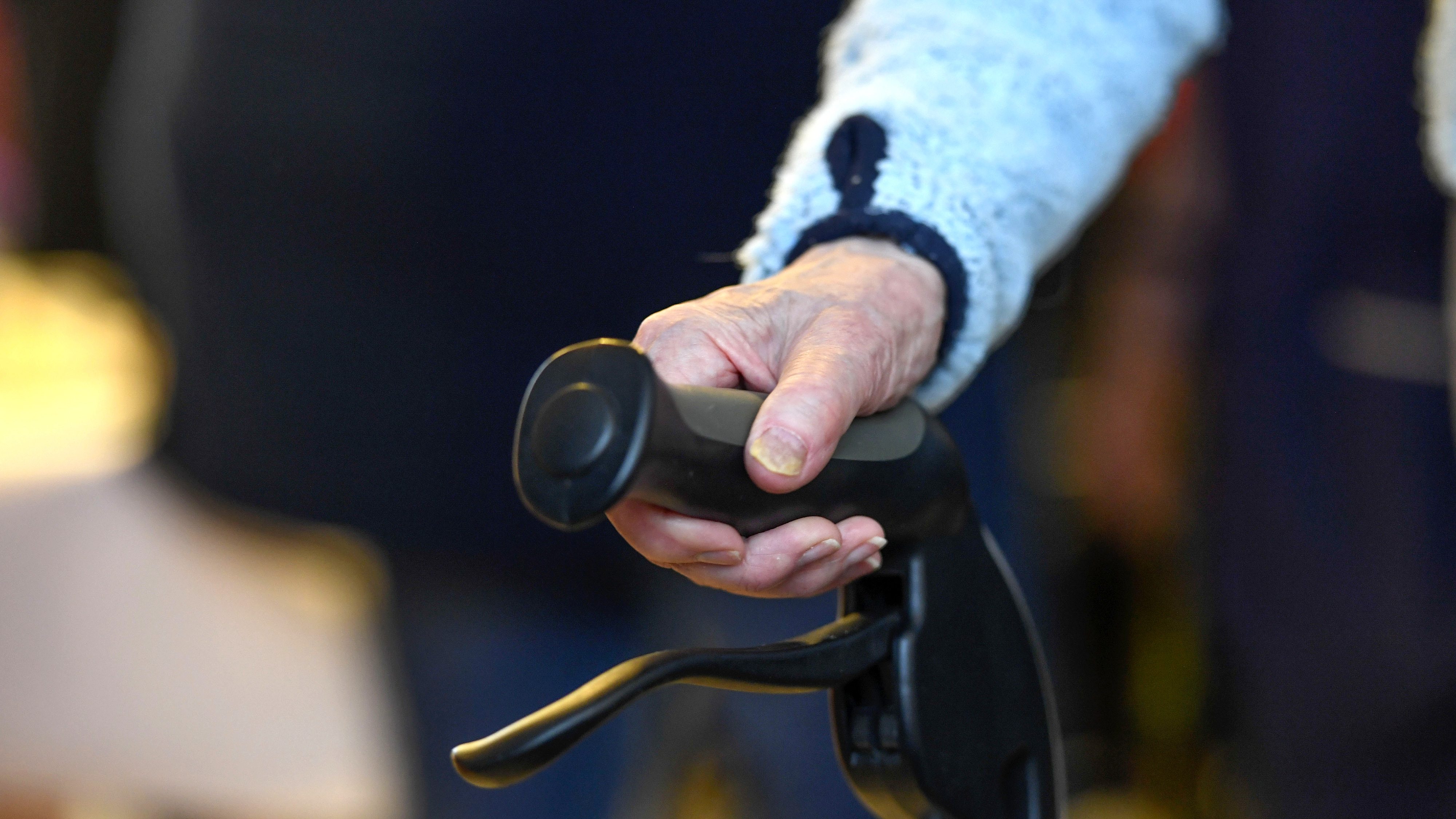 En äldre persons hand håller i handtaget på en rollator eller gåhjälpmedel, med suddig bakgrund.