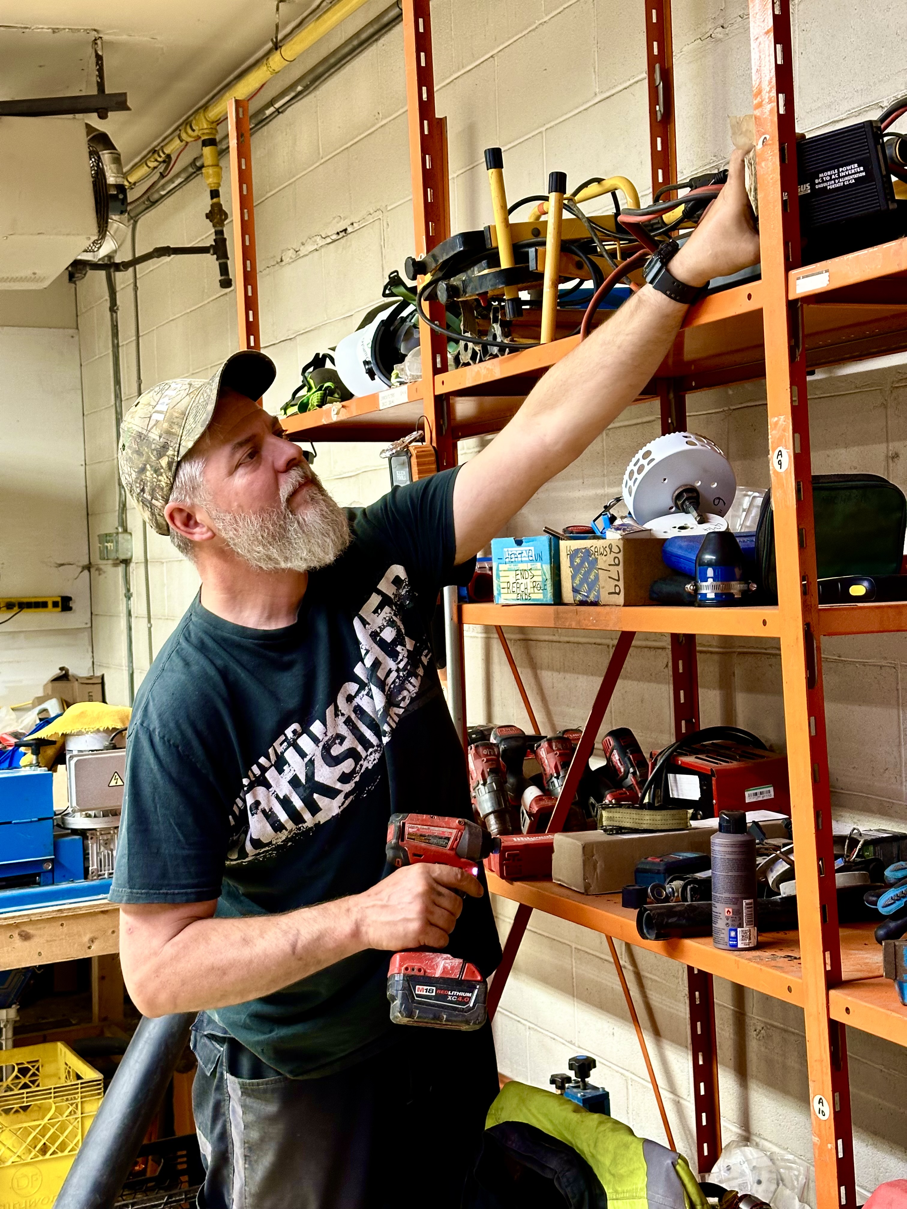 En man med grått skägg och keps håller en borrmaskin och sträcker sig mot en hylla i en verkstad med verktyg och utrustning.