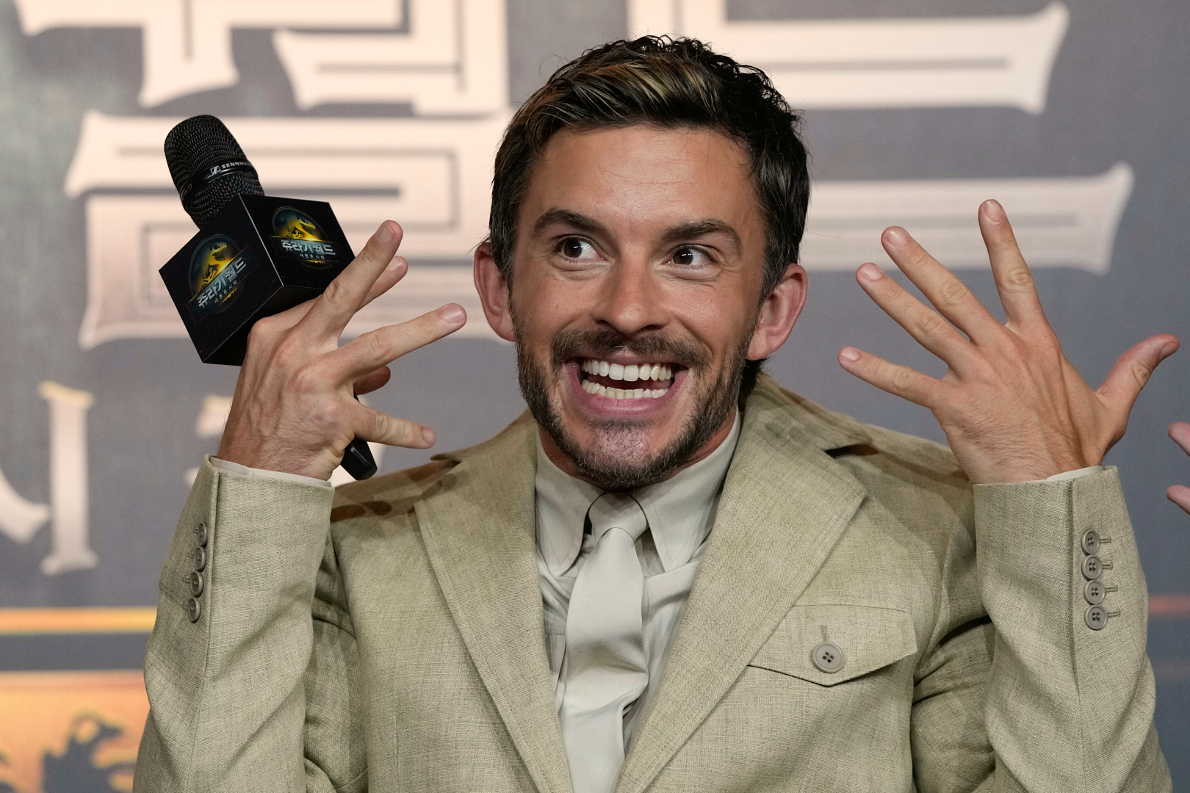 En man i beige kostym ler och gestikulerar med båda händerna medan han håller en mikrofon under ett evenemang.