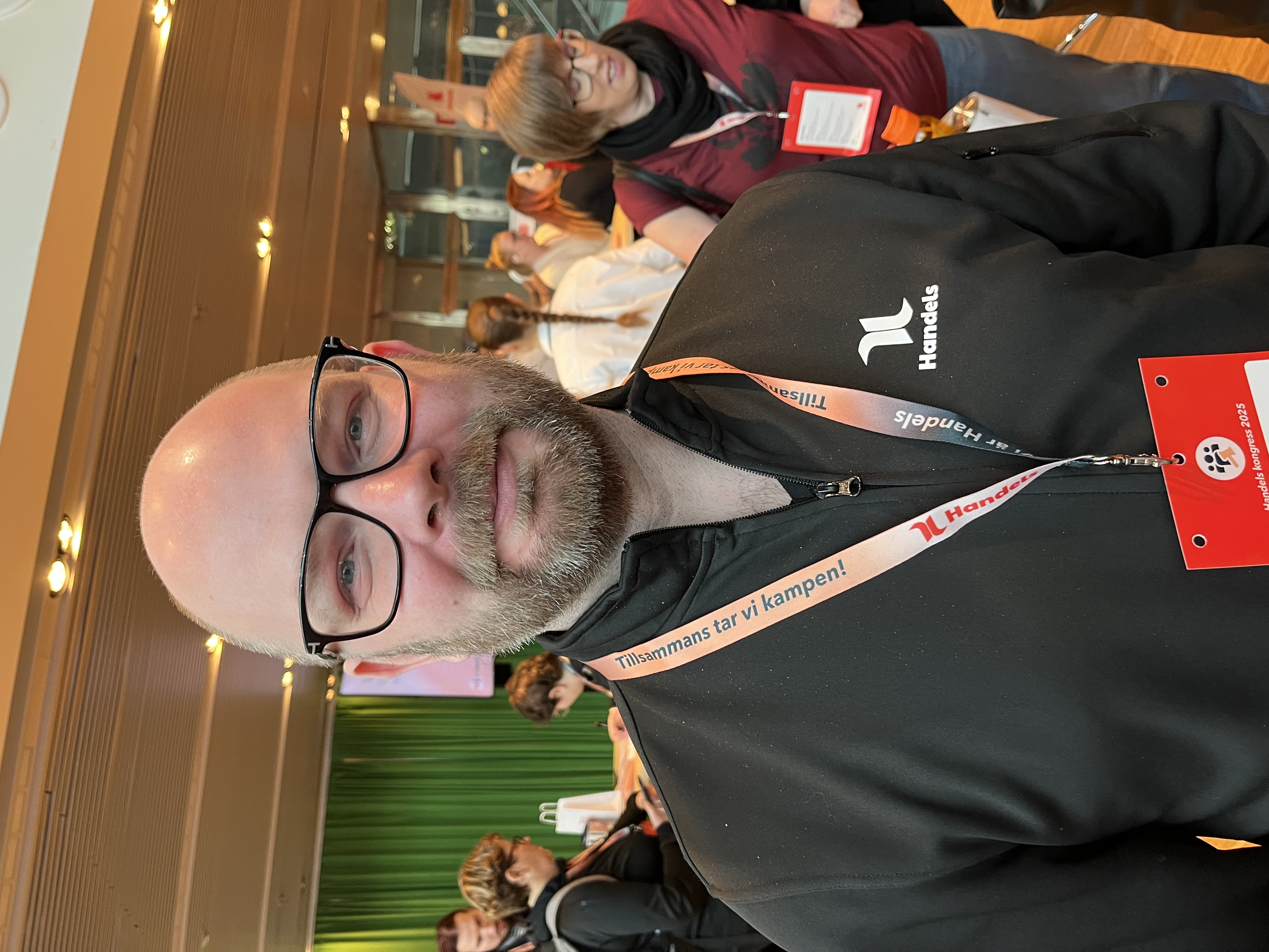 En man med glasögon och skägg bär en svart Handels-jacka och konferensbricka, står inomhus med andra personer i bakgrunden.