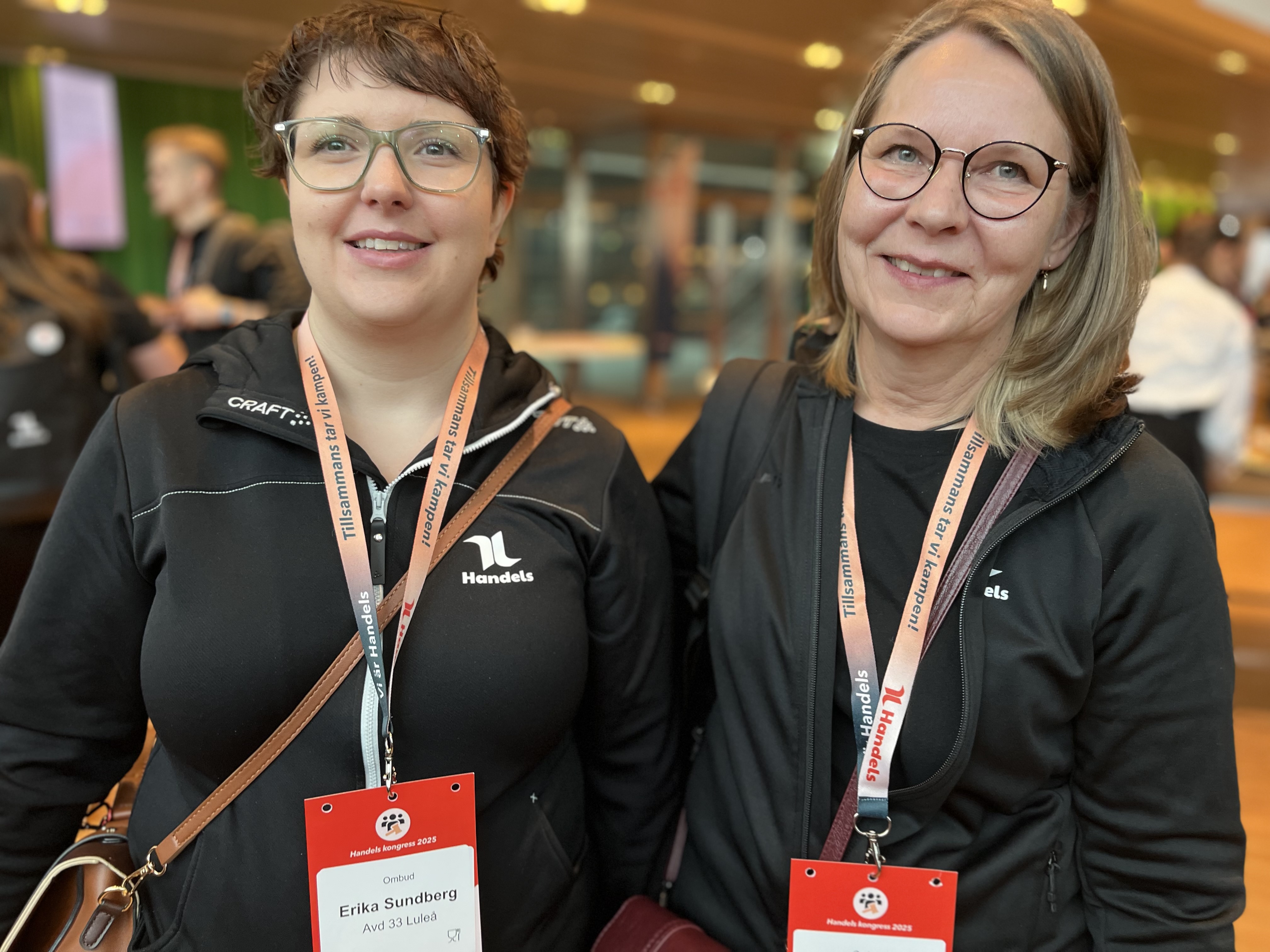 Två kvinnor med glasögon och svarta jackor står inomhus med konferensband och namnskyltar runt halsen. Bakgrunden är suddig.