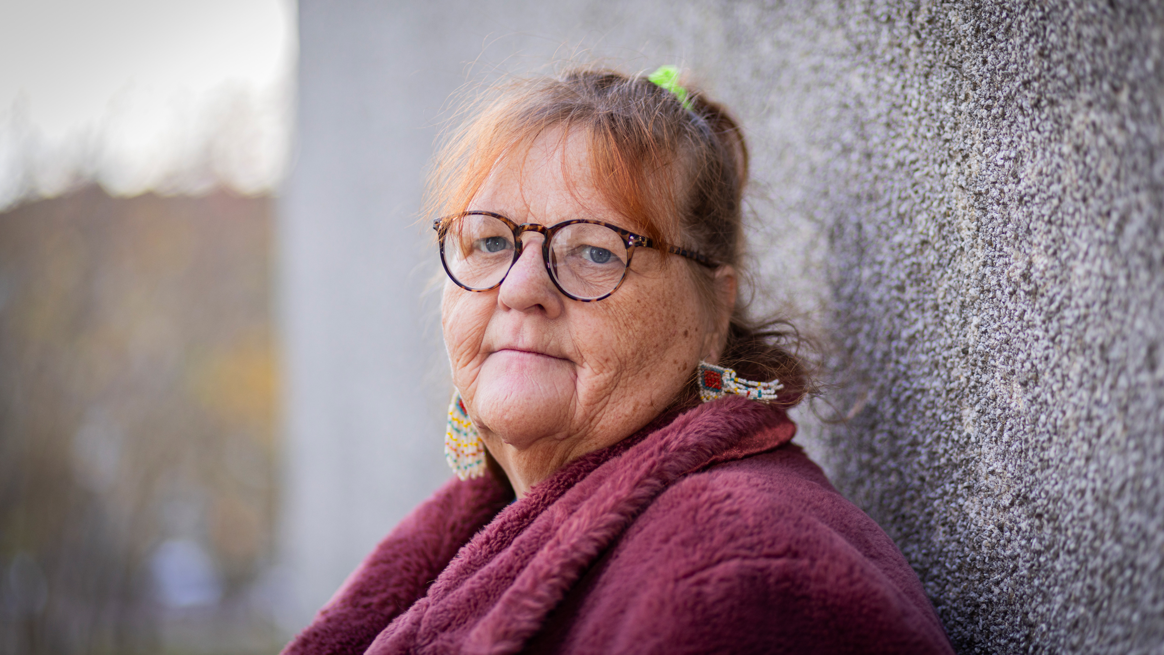 En äldre kvinna med glasögon och rött hår, i en mörkröd kappa, lutar sig mot en vägg utomhus och tittar mot kameran.