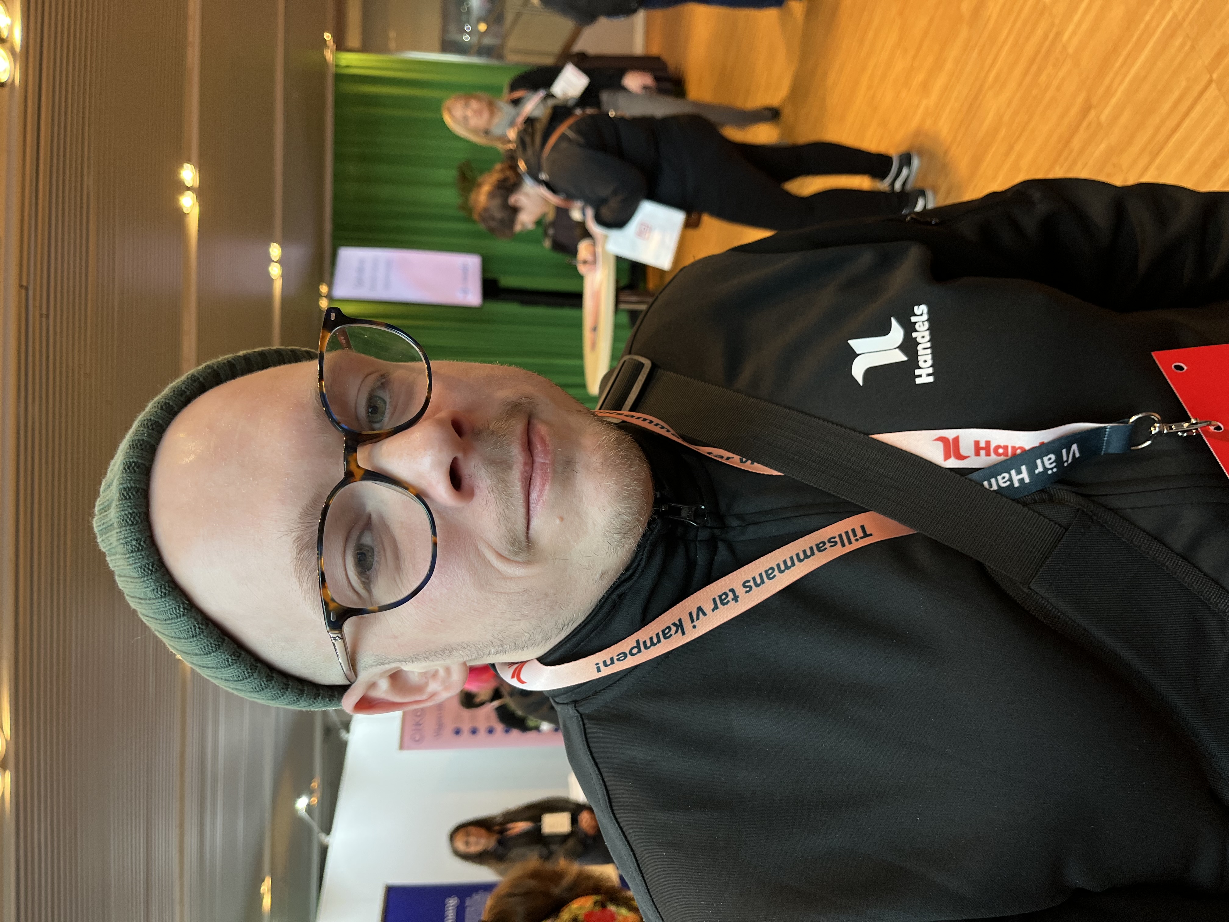 En person med glasögon, grön mössa och svart Handelsjacka står inomhus på ett evenemang. Flera personer och montrar syns i bakgrunden.