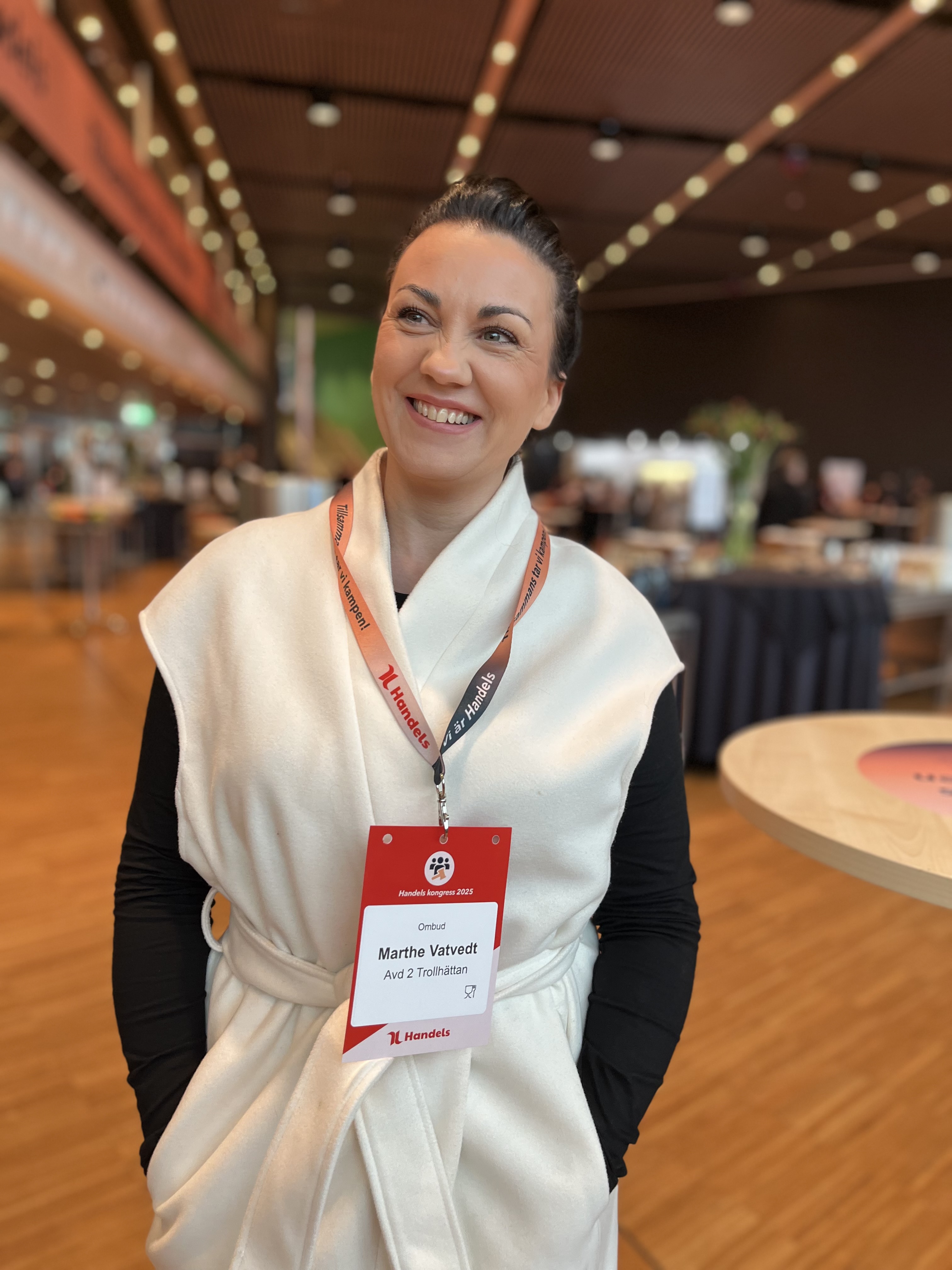 En kvinna med vitt väst och svart topp står inomhus på ett evenemang med en namnskylt där det står "Martha Viñvet".