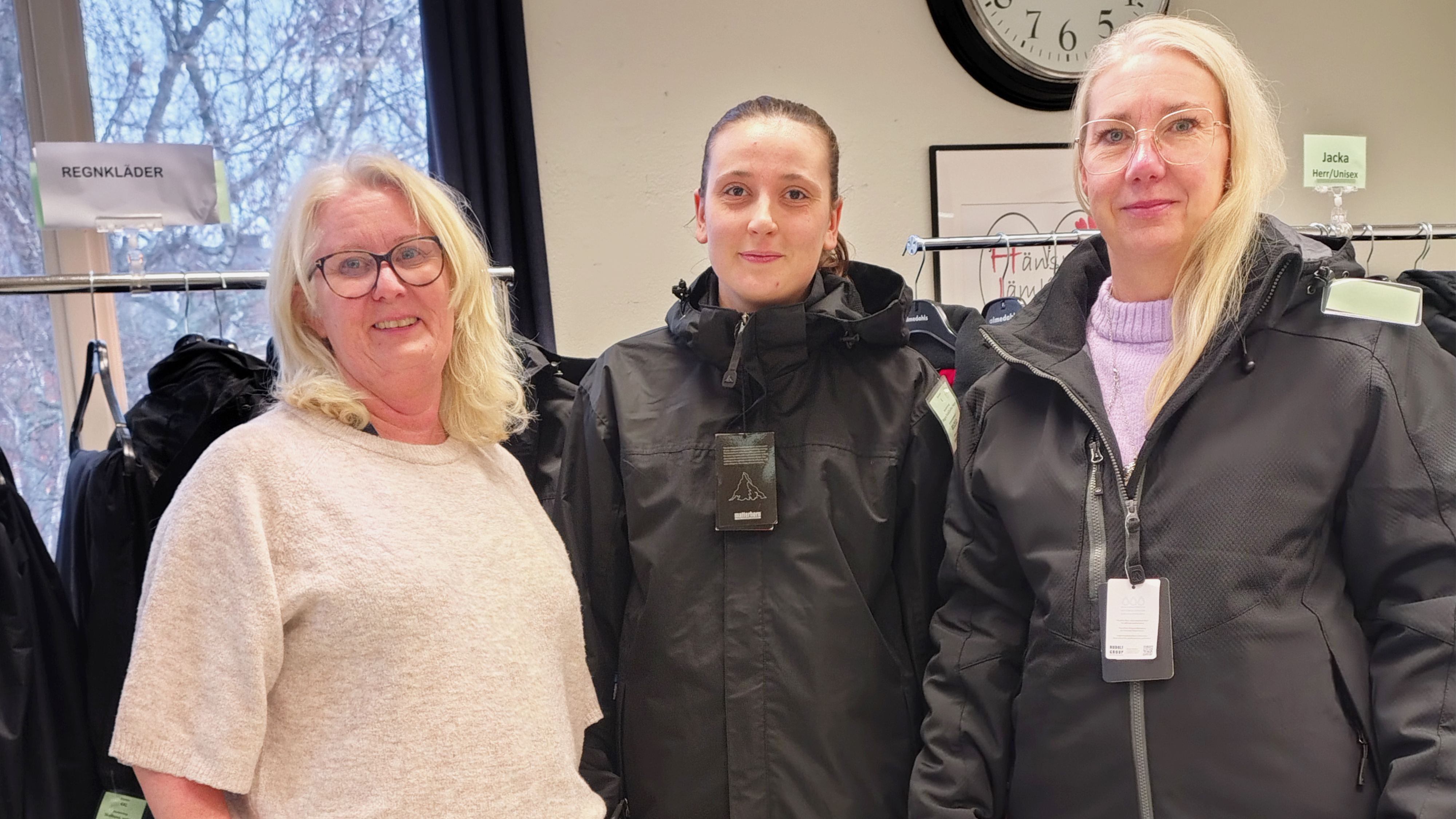 Tre kvinnor står inomhus, två har svarta jackor med lappar och en har en ljus tröja. Klädställ och klocka i bakgrunden.
