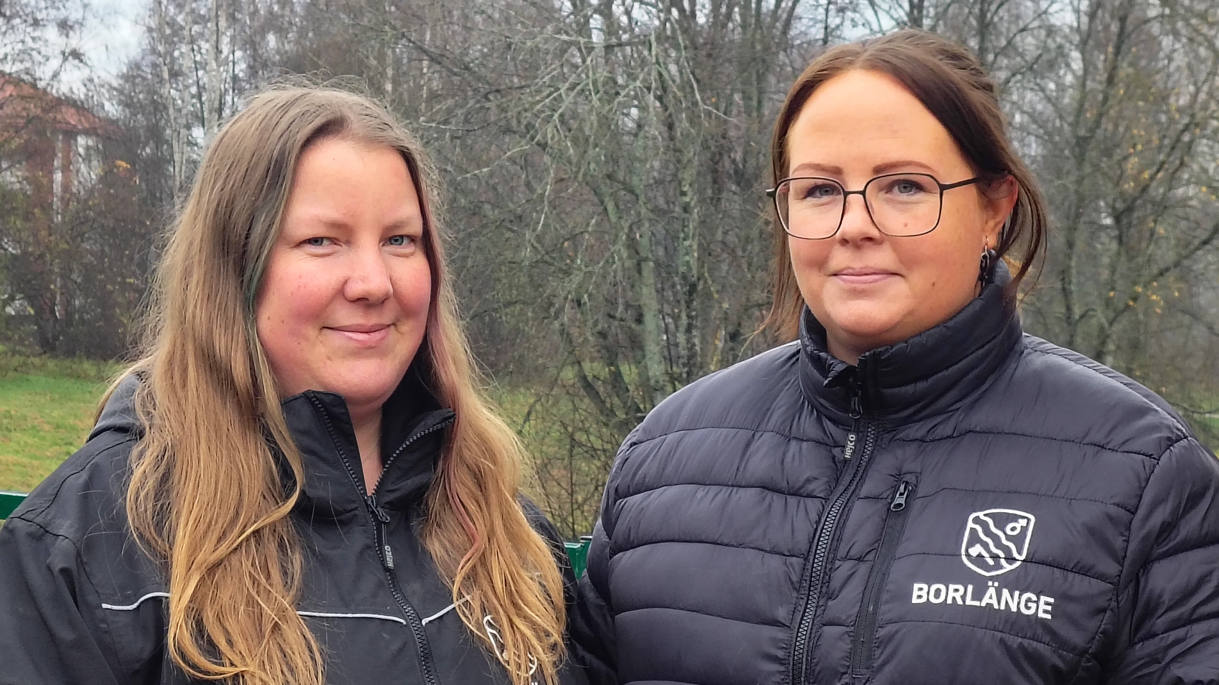Två kvinnor står utomhus och har svarta jackor med "BORLÄNGE"-logga; träd och molnig himmel syns i bakgrunden.