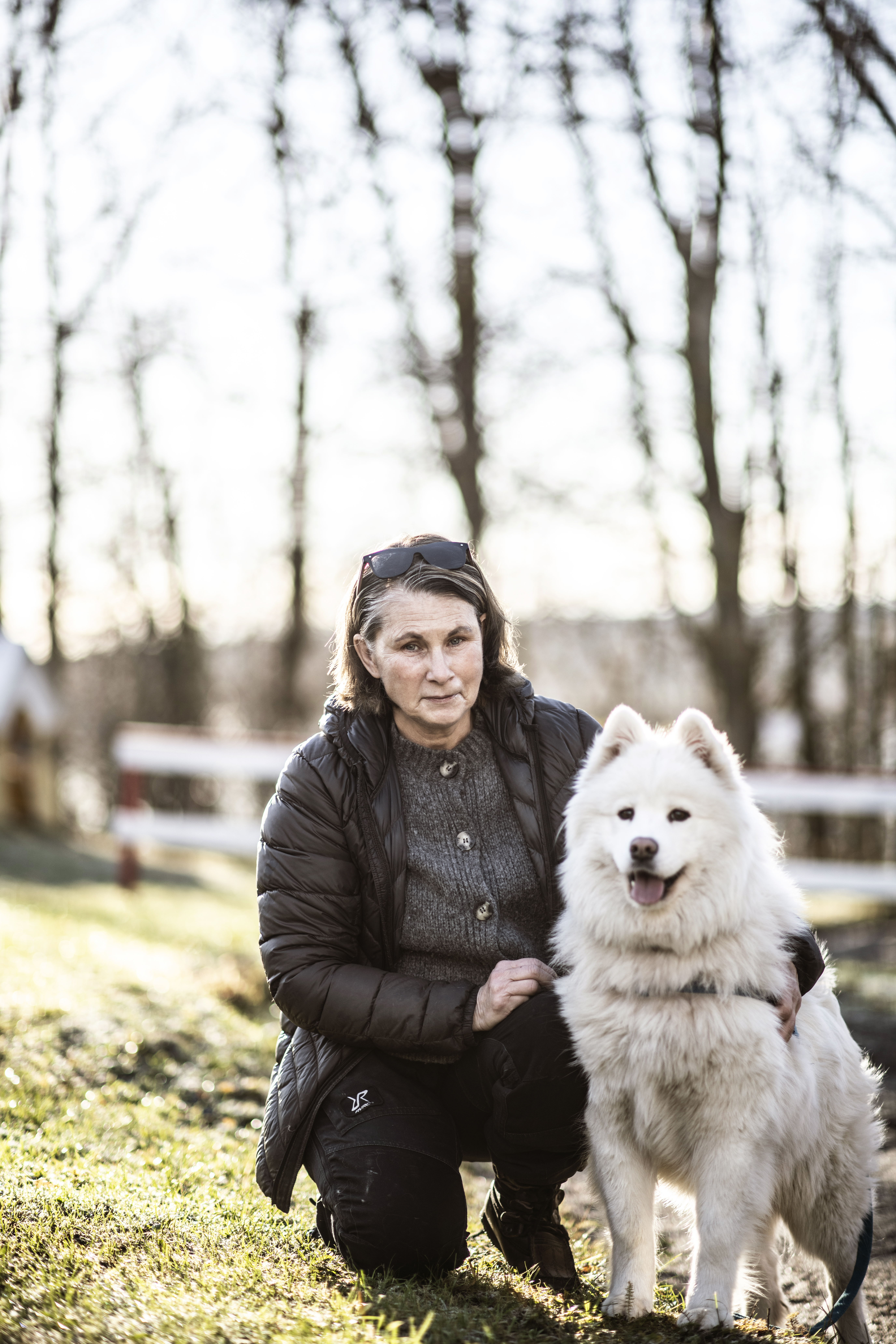 En kvinna med mörk jacka knäböjer på gräs bredvid en vit hund utomhus med träd utan löv i bakgrunden.