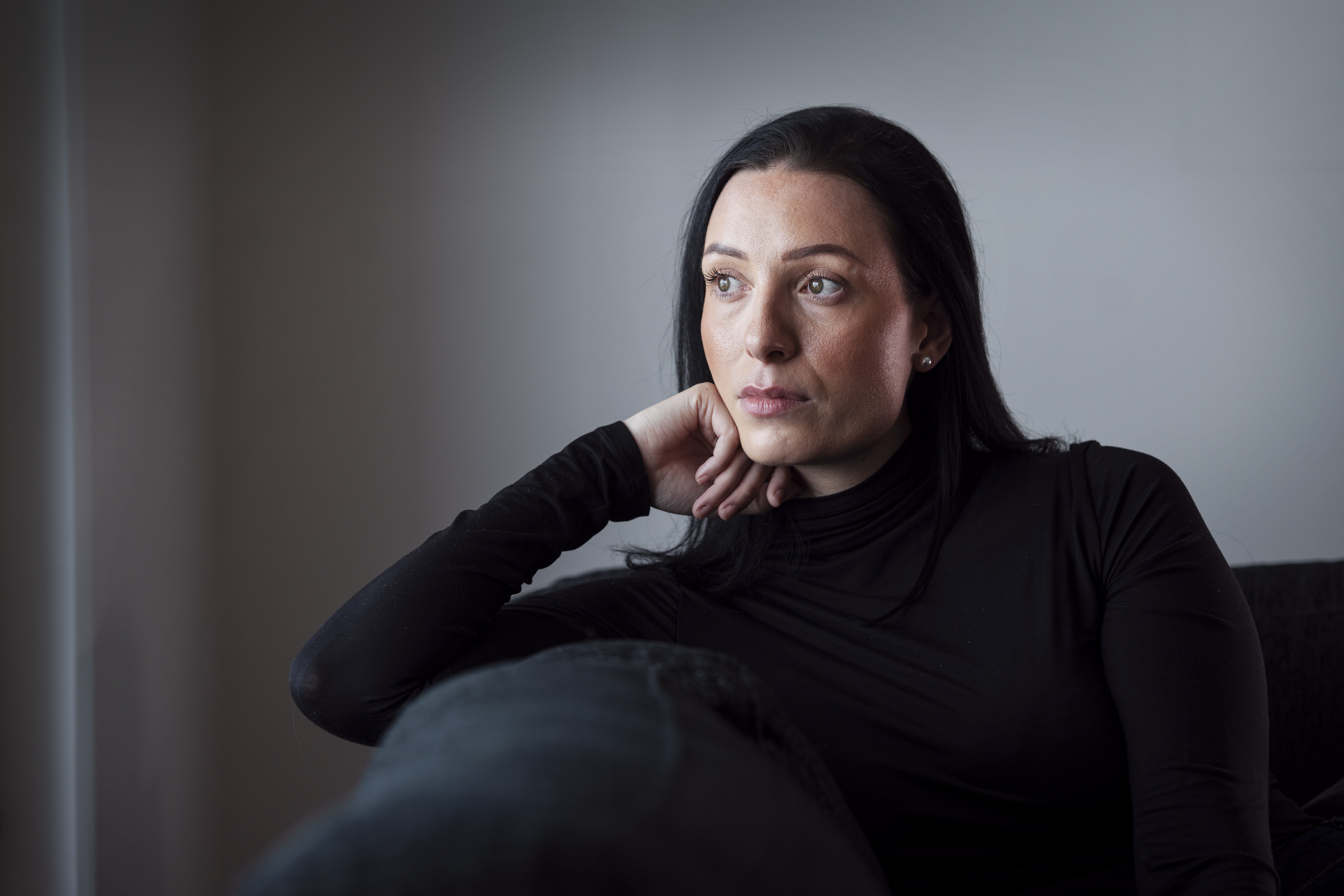 Lina med långt mörkt hår och svart polotröja sitter inomhus med huvudet vilande mot händerna och blicken i fjärran.