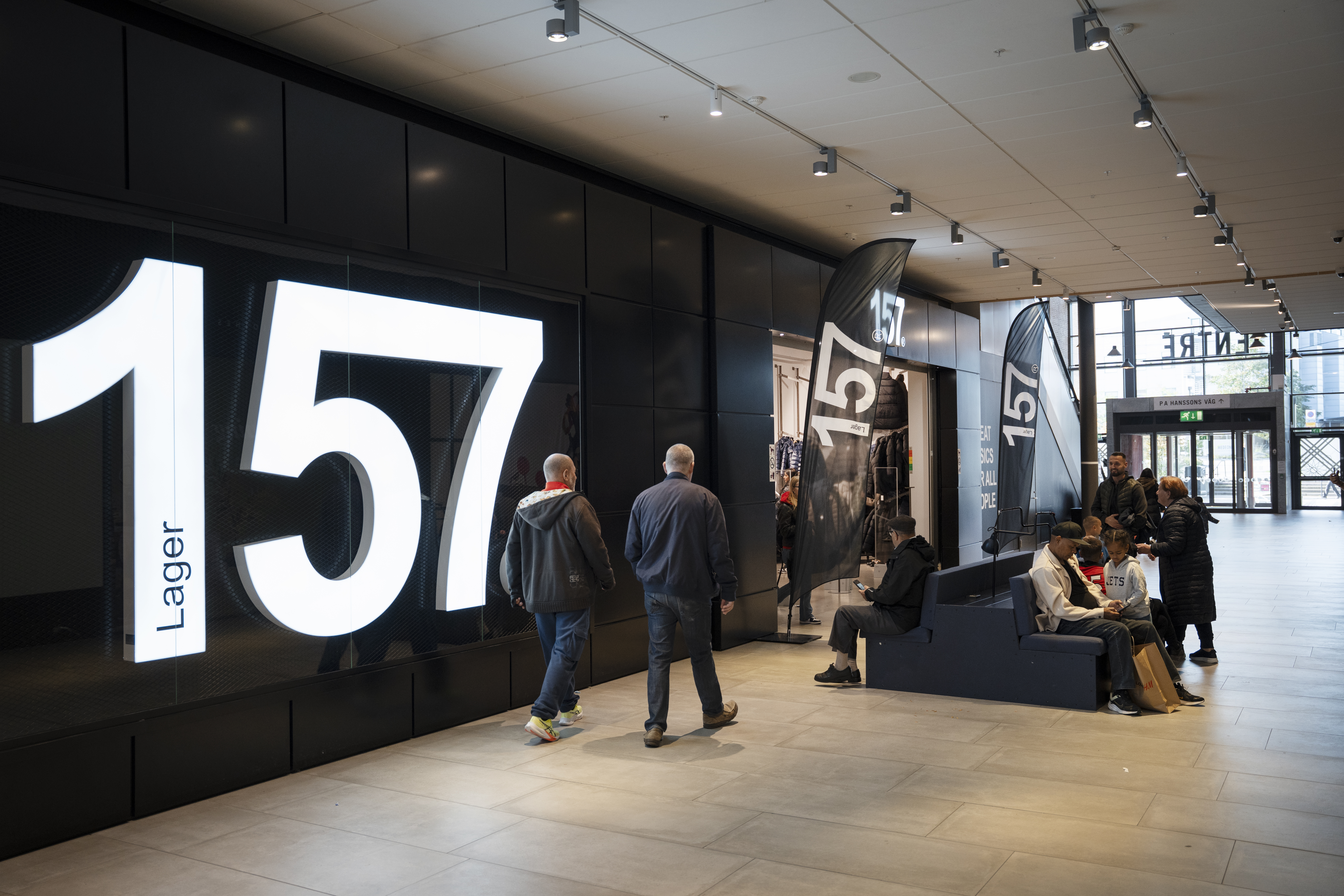 Personer går och sitter utanför en butik med en upplyst skylt där det står "Lager 157" på väggen.