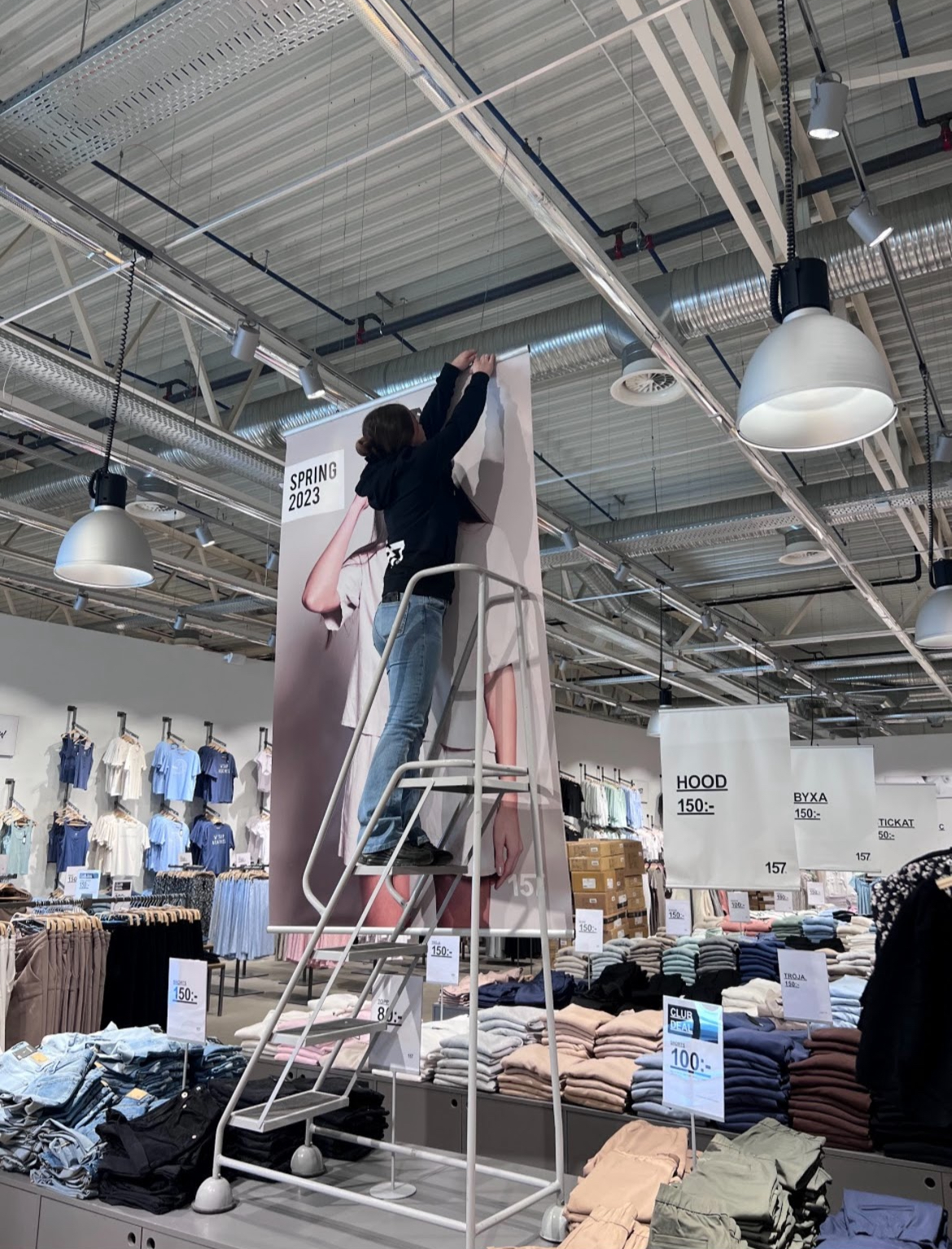 En person står på en stege på ett bord och justerar en stor modeaffisch i taket i en butik.