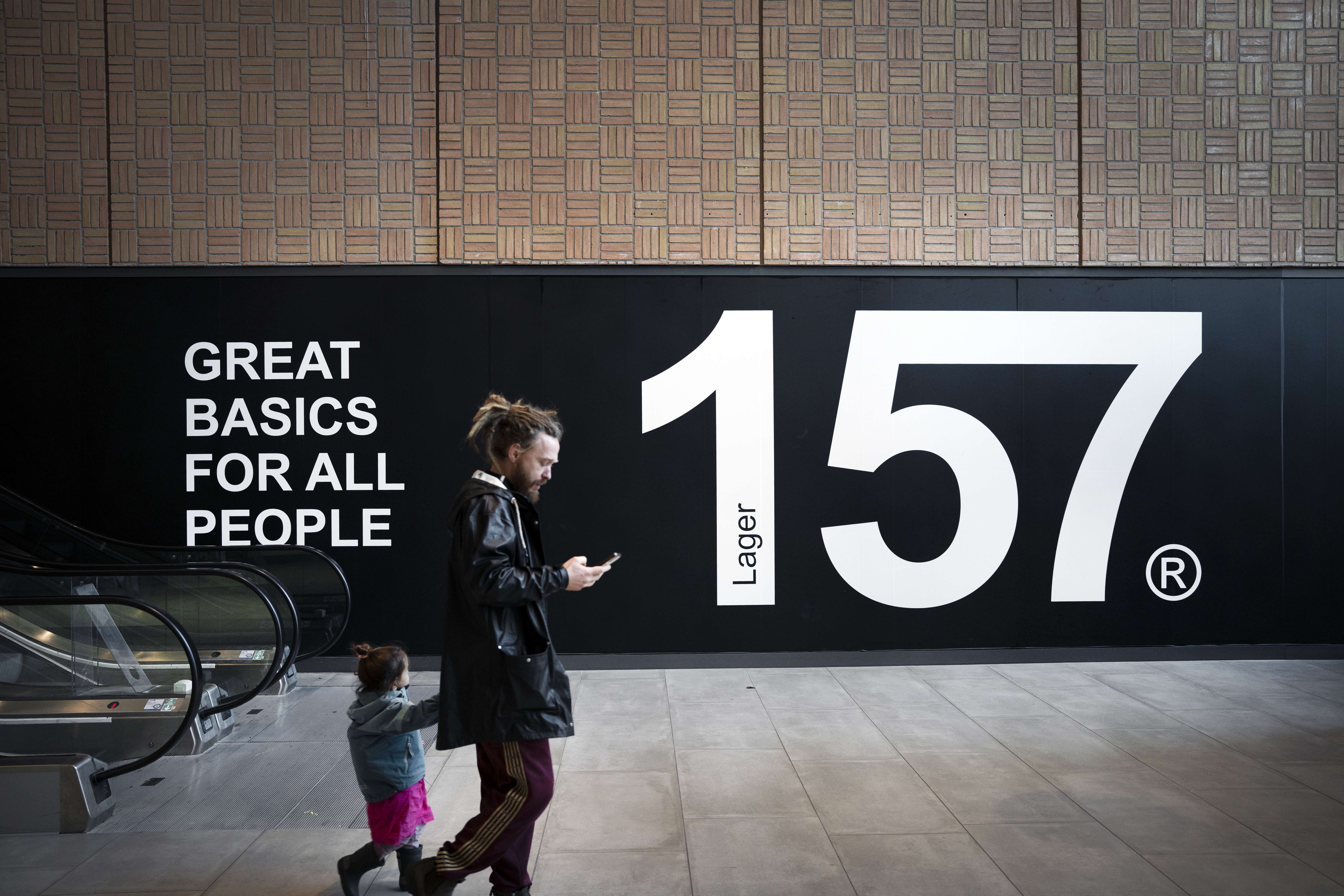 En man och ett barn går inomhus förbi en svart vägg med vit text där det står ”157” och texten ”Great basics for all people”.