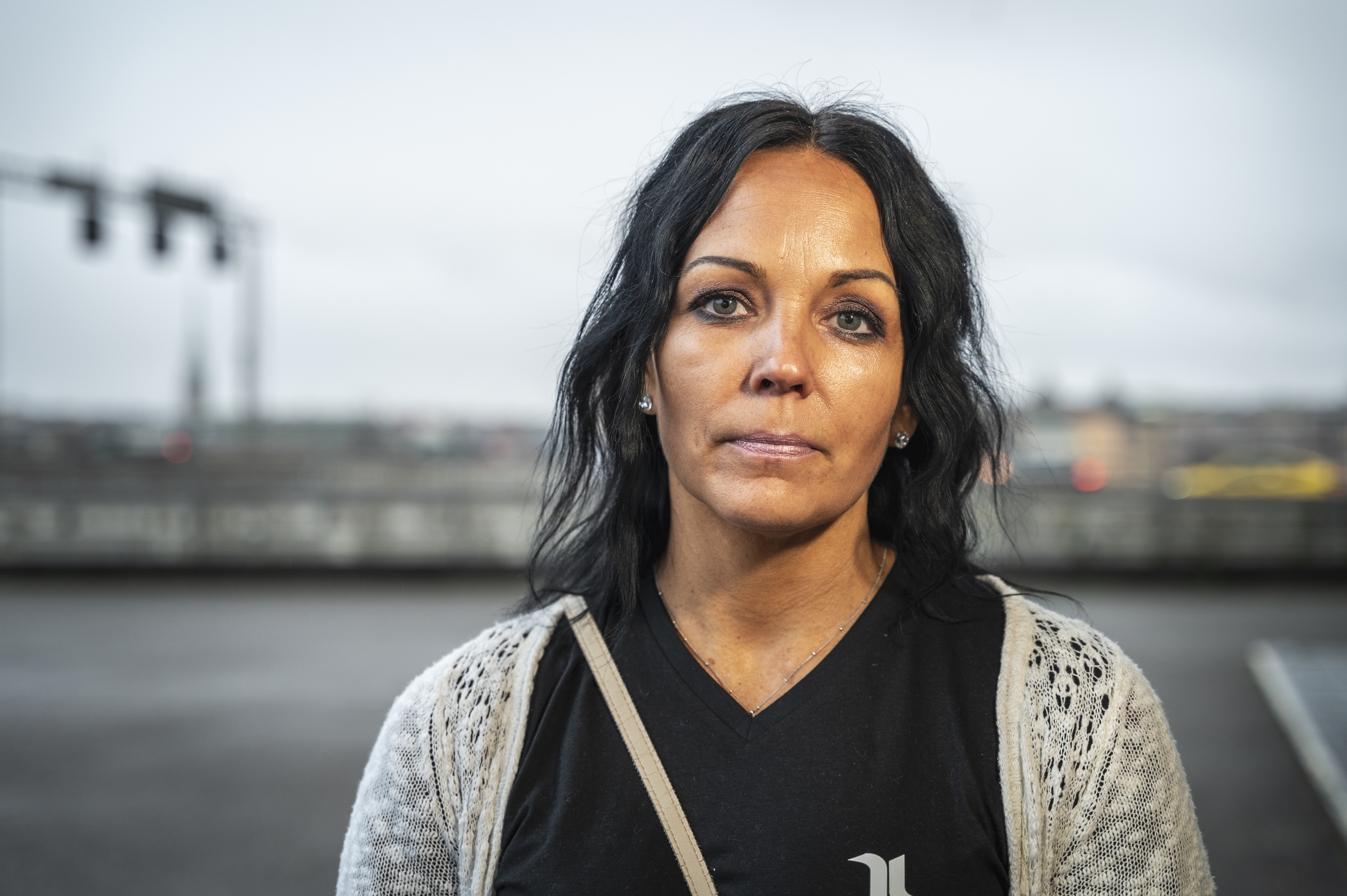 En kvinna med långt mörkt hår och neutralt ansiktsuttryck står utomhus framför en suddig stadsbakgrund.