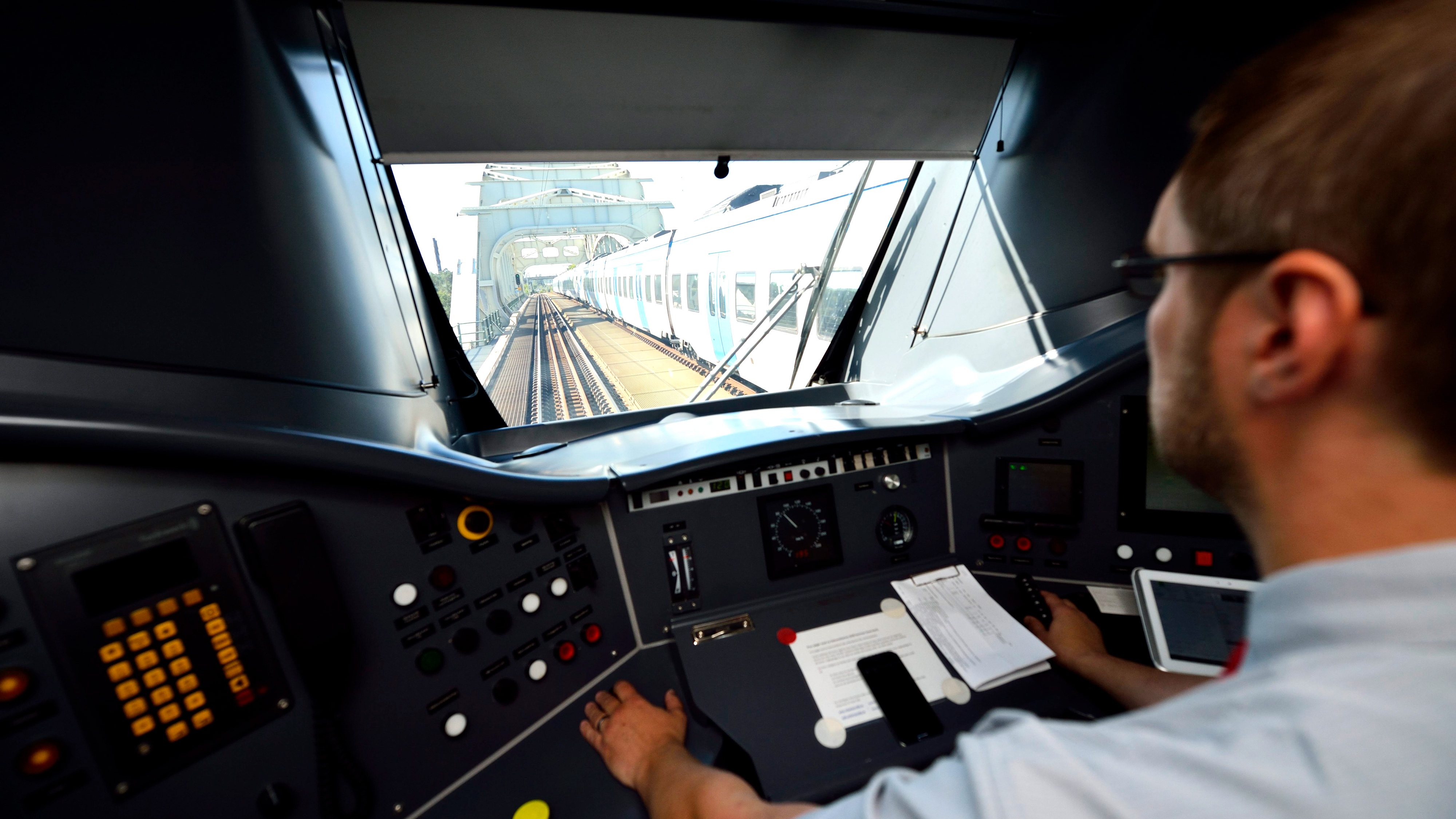 Tågförare sitter i förarhytten och styr ett tåg på spår mot en bro med ett annat tåg synligt längre fram.