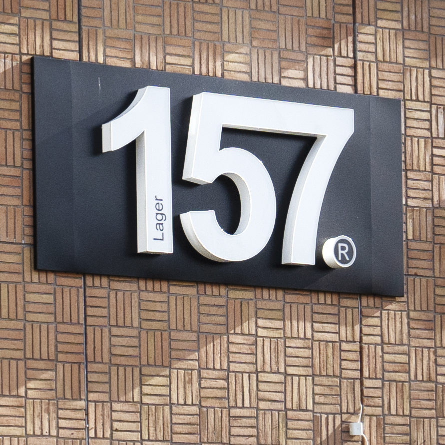 Stor vit skylt med texten "157® Lager" sitter på en brun mur med mönster.