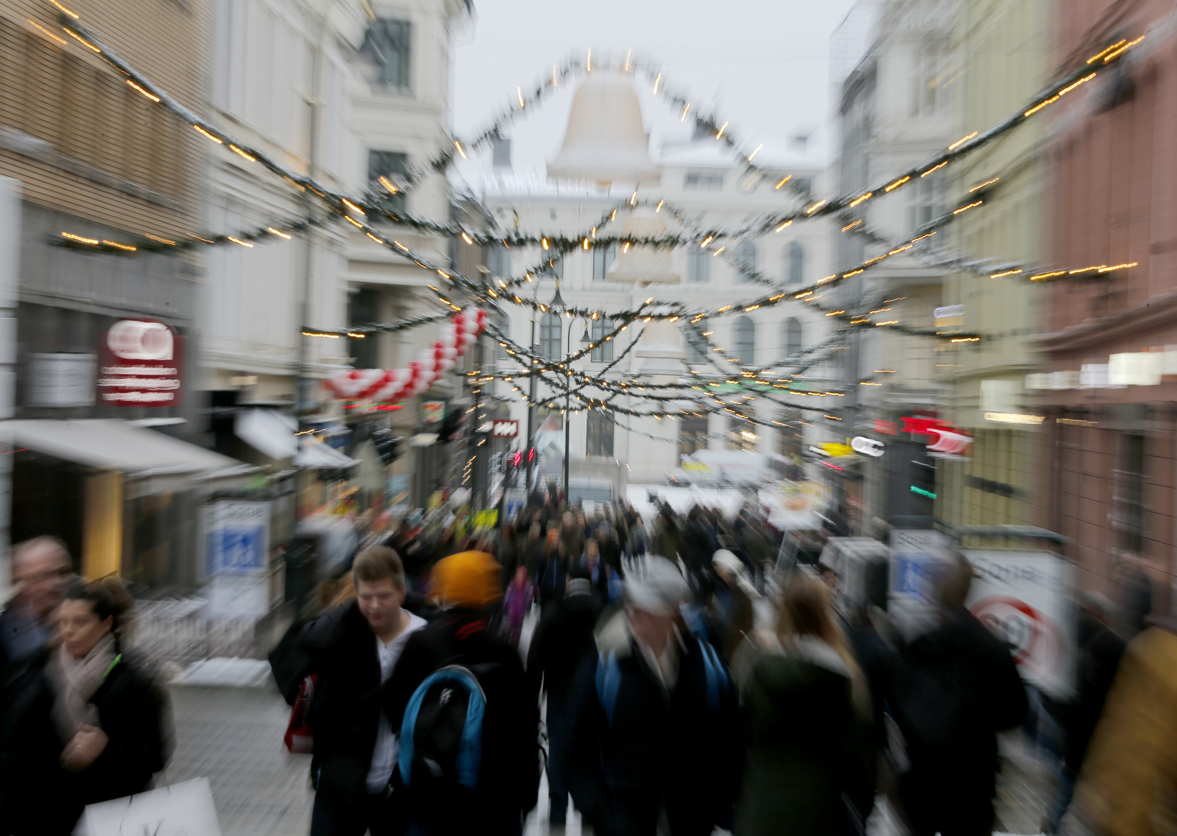 En stadsgata med människor som går, dekorerad med ljusslingor och julgirlanger, är suddig och oskarp.