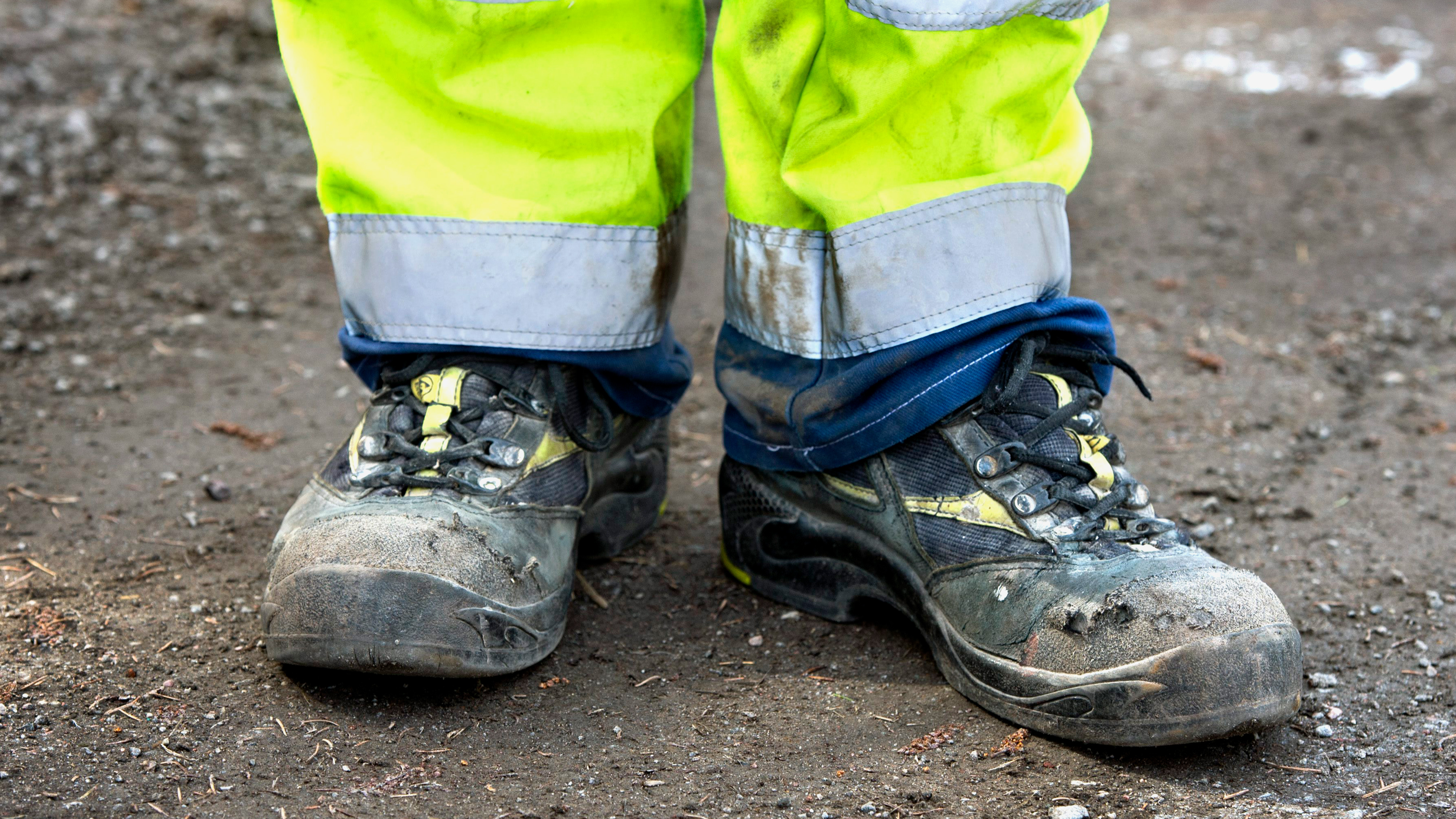 En person med svarta arbetskor och gula varselbyxor står på en yta med grus och jord.
