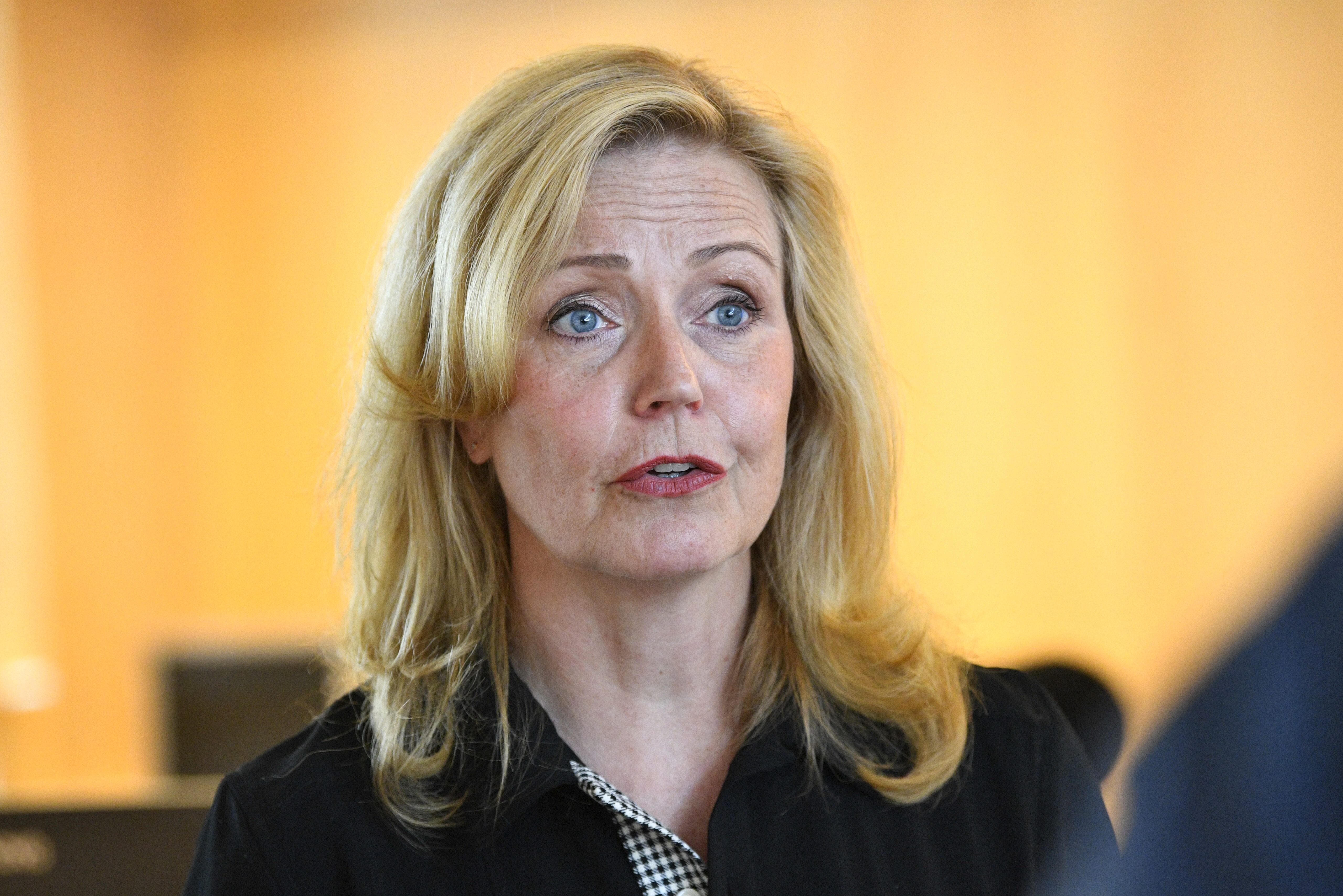 En kvinna med blont hår och svart topp tittar lite åt sidan och pratar inomhus. Bakgrunden är suddig.