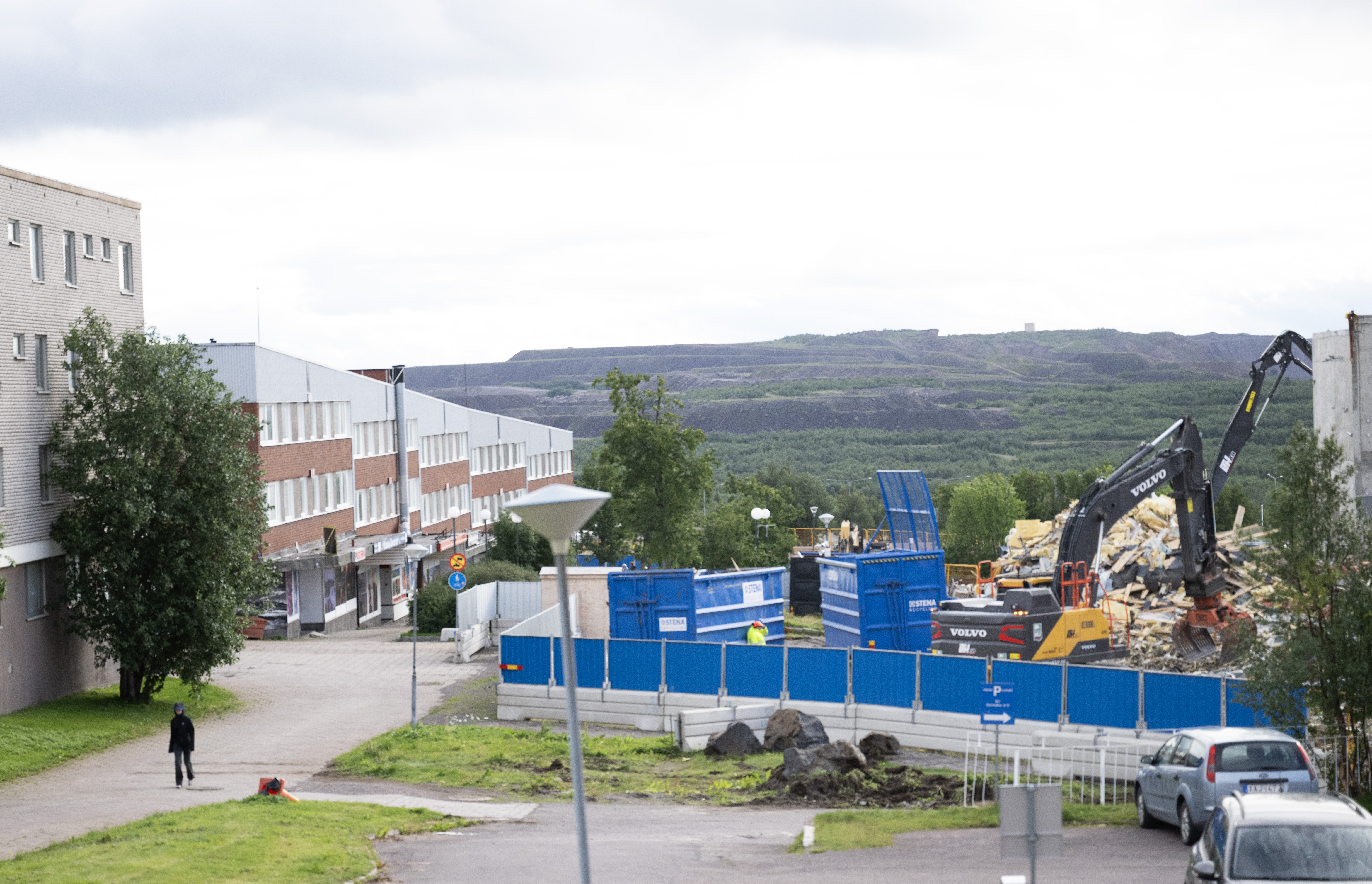 Byggarbetsplats med grävmaskiner och blå containrar bredvid en byggnad, inhägnad med blå staket; en person går på en stig, kullar syns i bakgrunden.