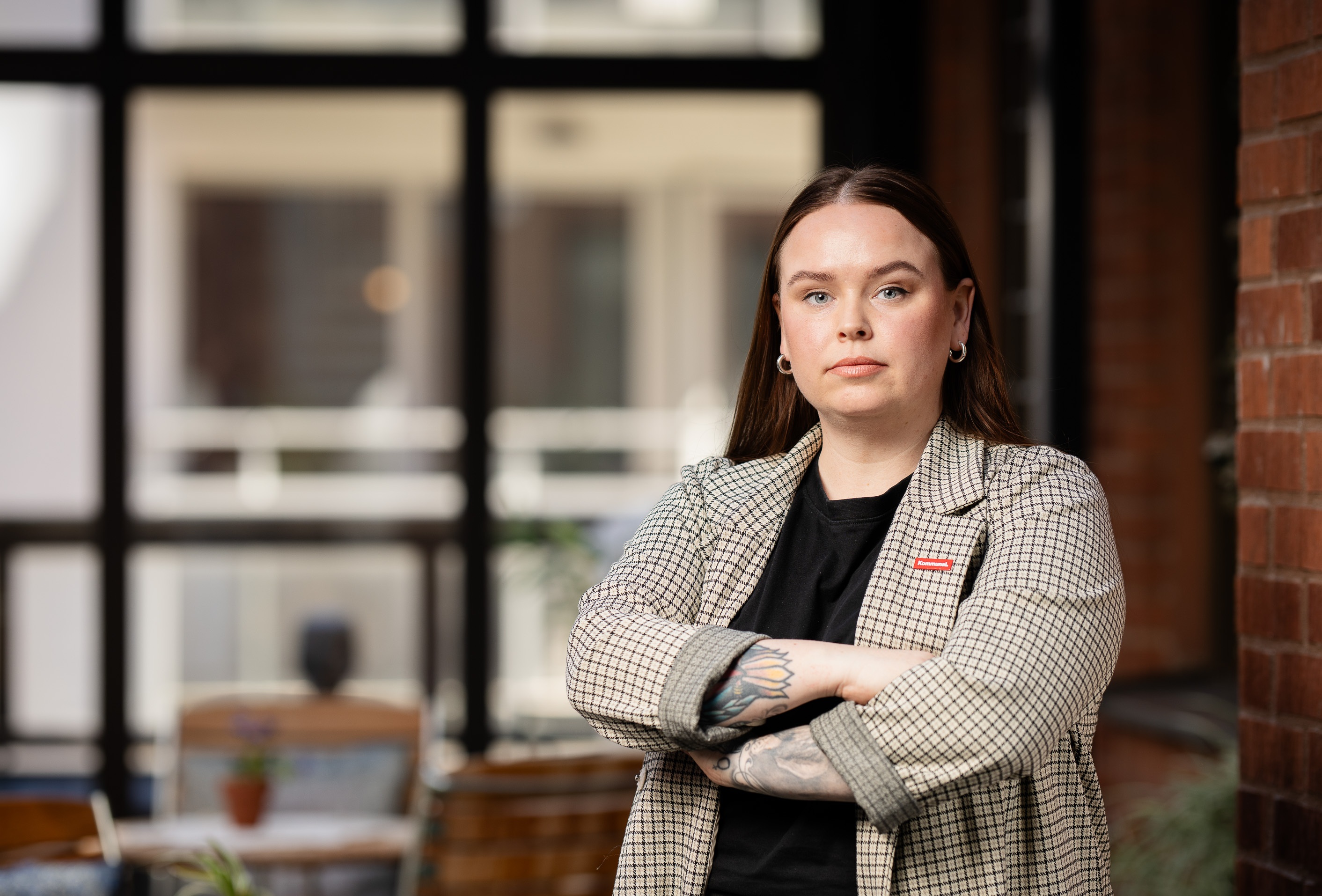 En kvinna med långt brunt hår och synliga tatueringar står inomhus med armarna i kors, iförd rutig kavaj och svart tröja framför stora fönster.