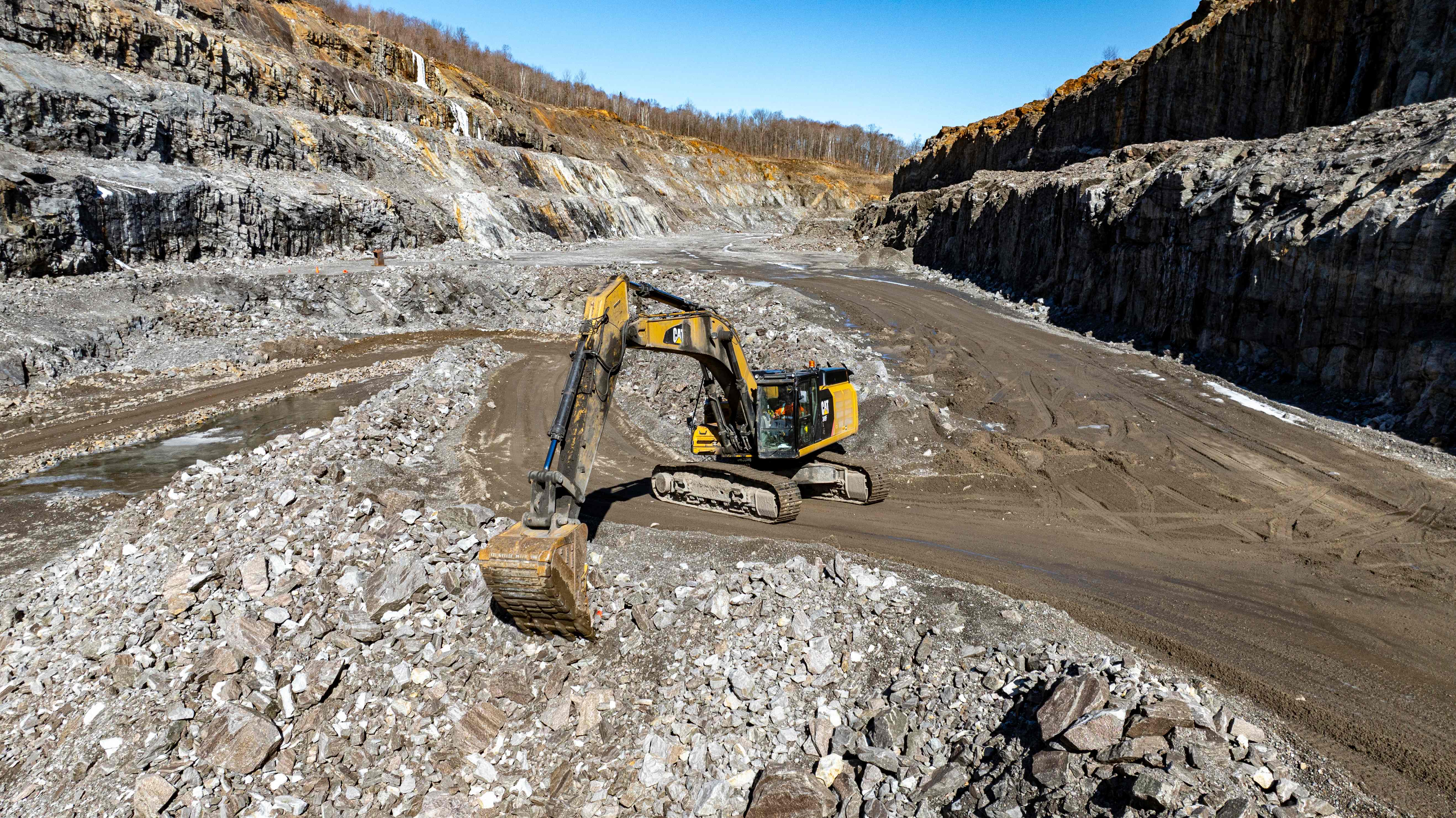 En gul grävmaskin arbetar i ett stort dagbrott omgiven av steniga väggar under en klar himmel.