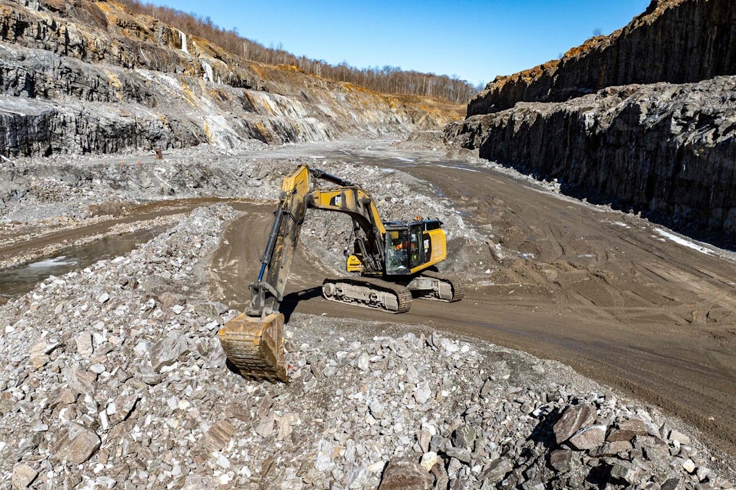 En gul grävmaskin arbetar i ett stort dagbrott omgiven av steniga väggar under en klar himmel.