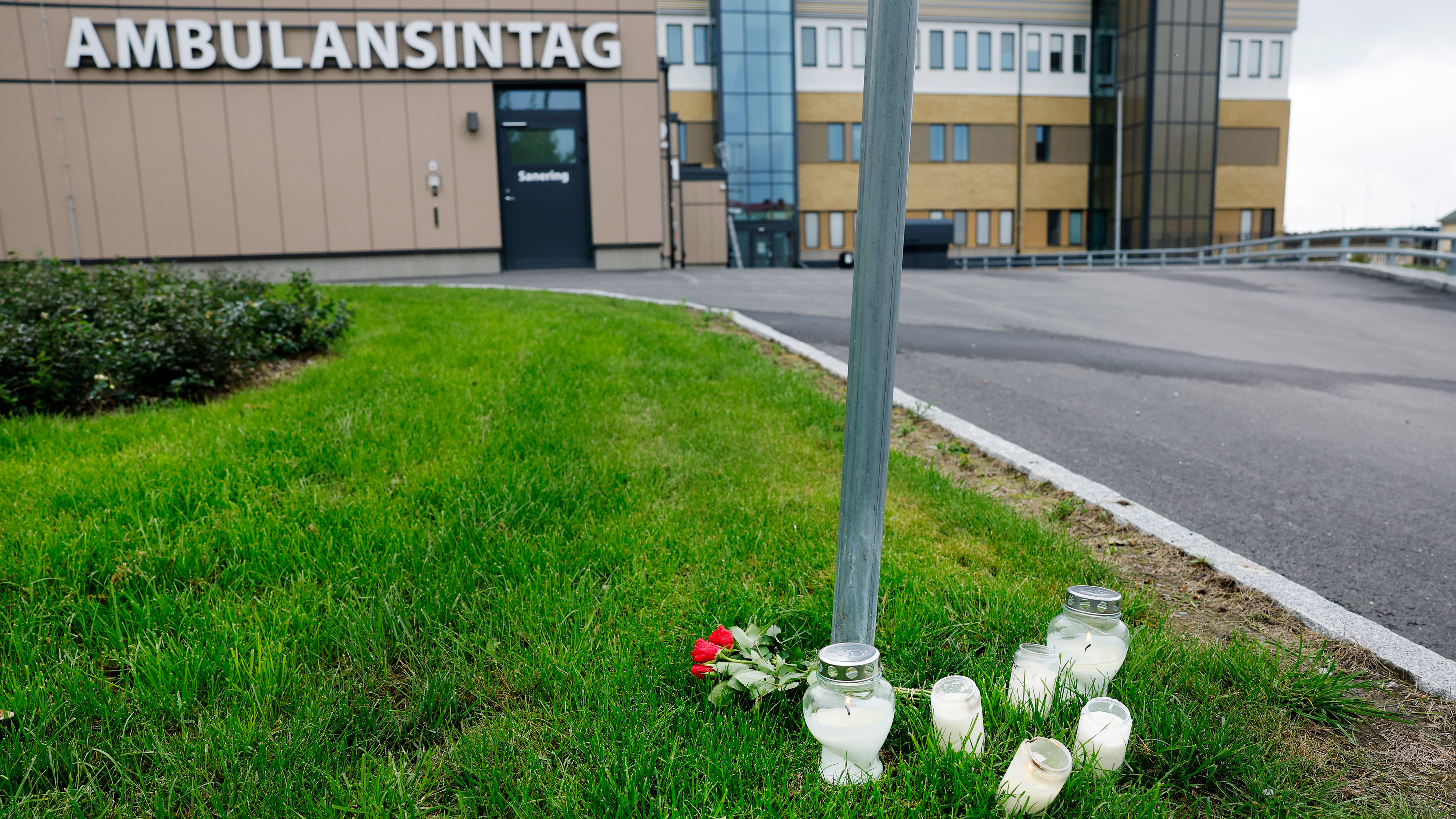 Flera tända ljus och röda rosor står på gräset vid en skylt utanför en byggnad märkt "AMBULANSINTAG".