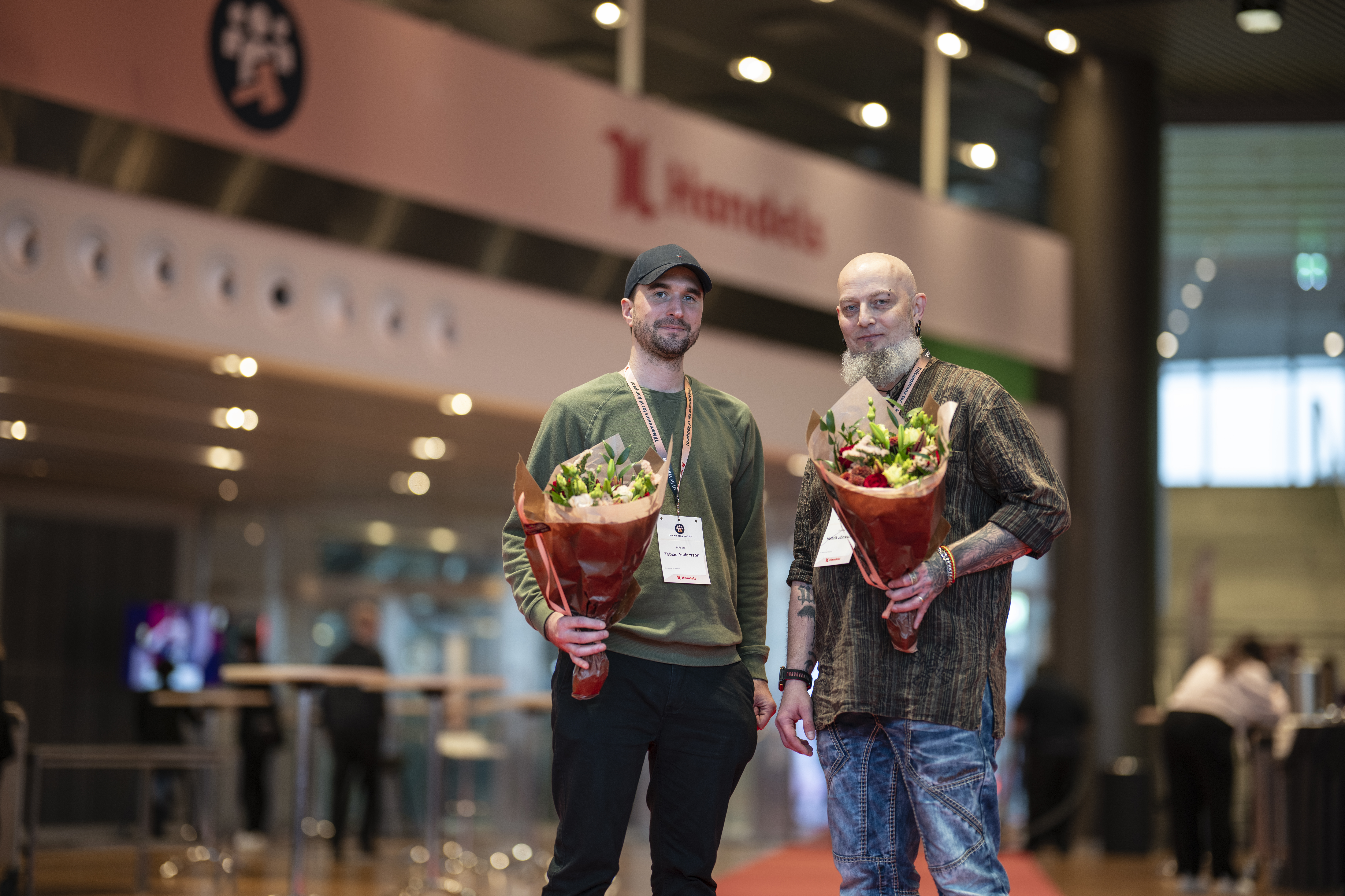 Två män står inomhus med stora blomsterbuketter och eventbrickor runt halsen, bord och skyltar syns i bakgrunden.