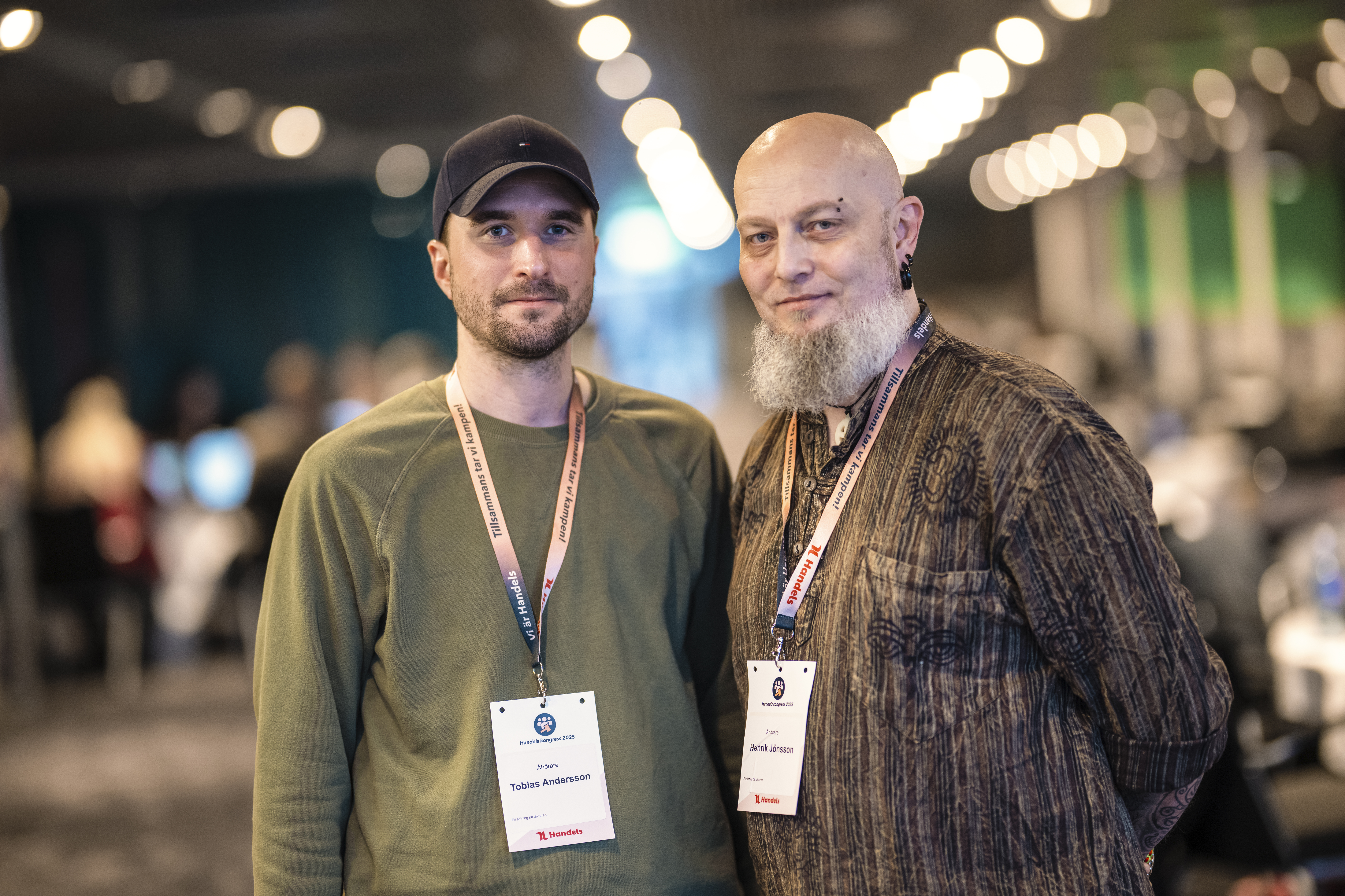 Två män med konferensbrickor står bredvid varandra inomhus; en har keps och grön tröja, den andra är skallig med vitt skägg och mönstrad skjorta. Bakgrunden är suddig.