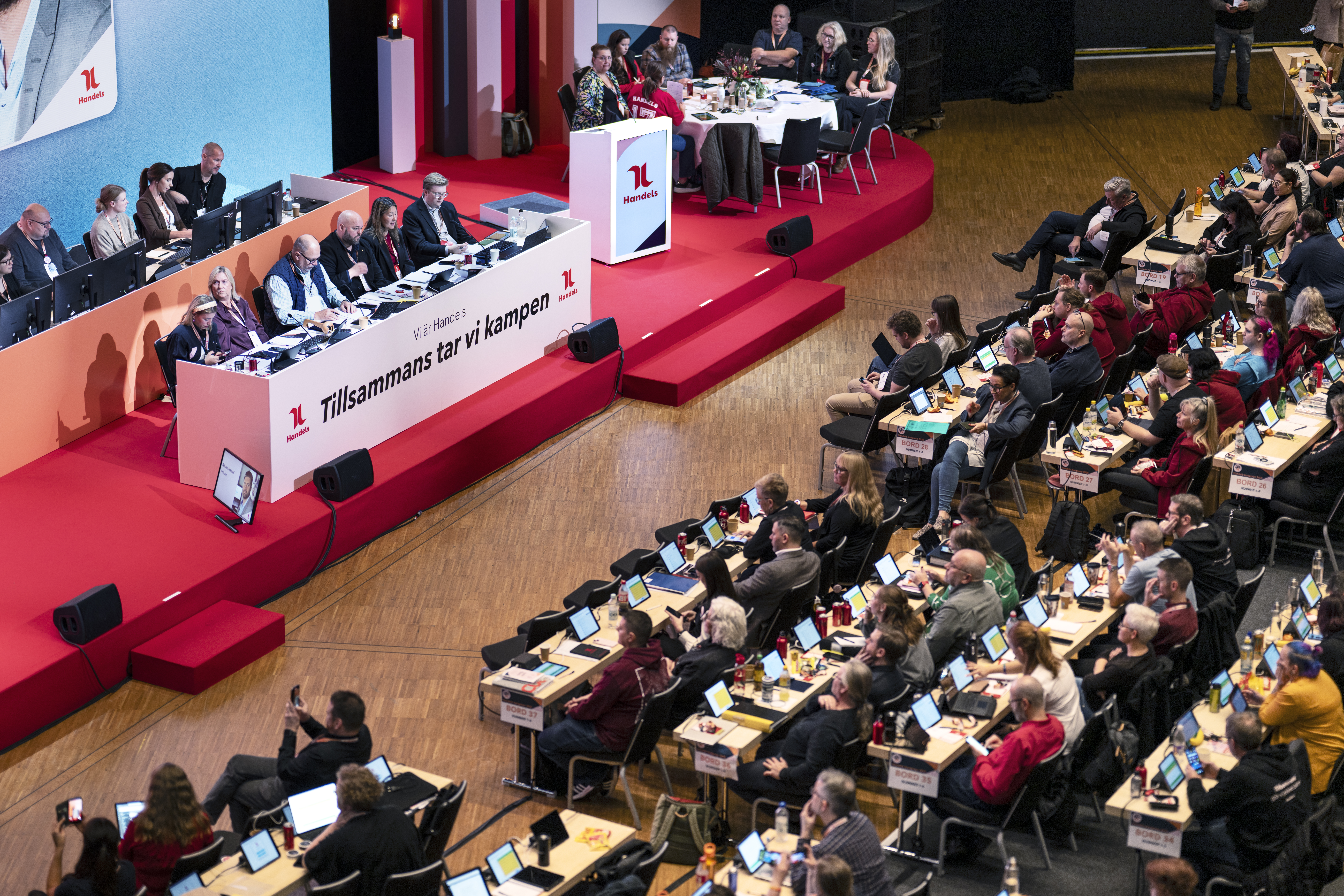 En kongressal uppifrån med rader av ombud som sitter vid skrivbord med surfplattor. På en röd scen sitter personer på två avlånga bord. Bordet längst fram har en vit framsida som det står Tillsammans tar vi kampen. På en talarstol står det Handels.