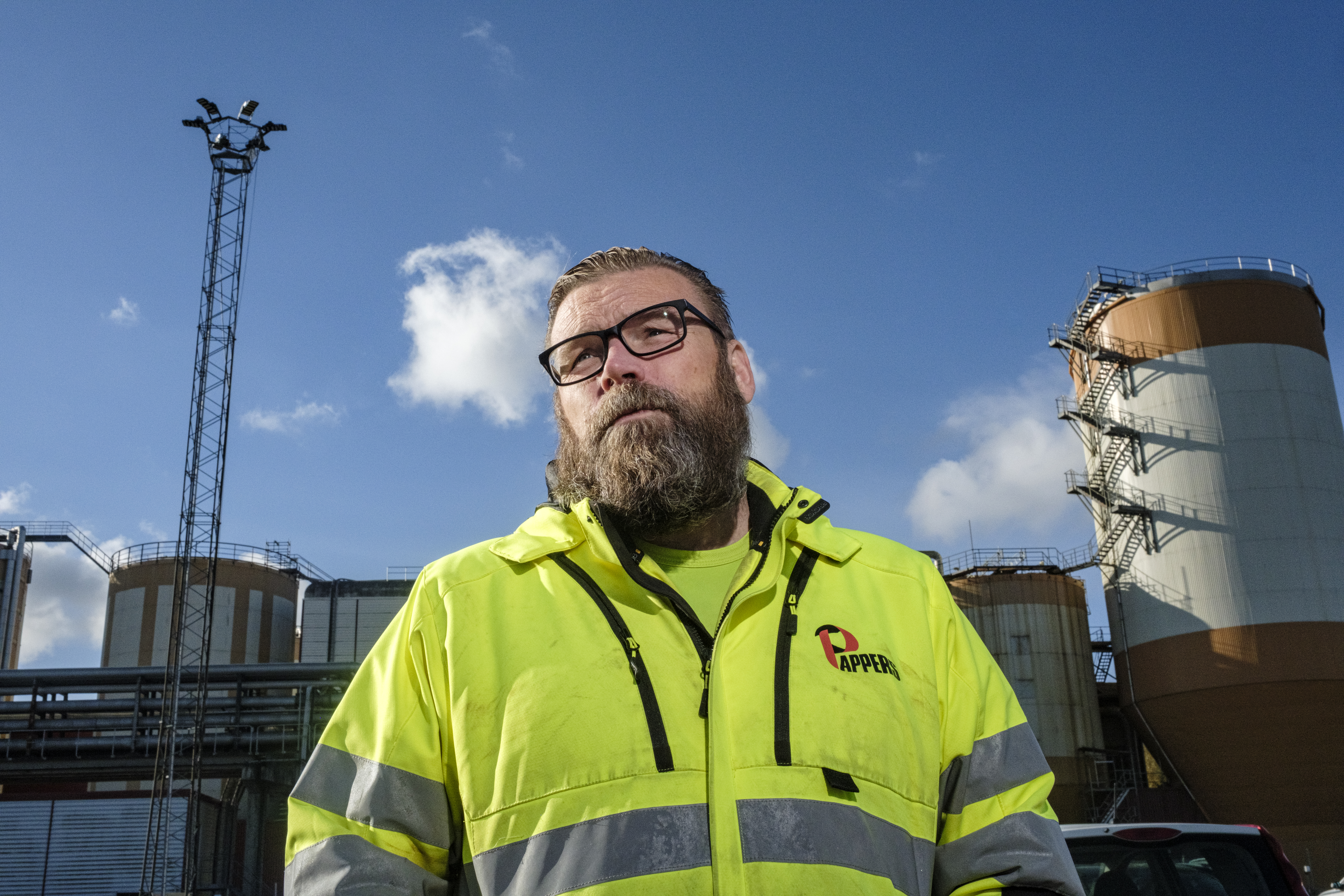 Lars G Karlsson med glasögon och varseljacka står utomhus framför industriella tankar och ett torn.